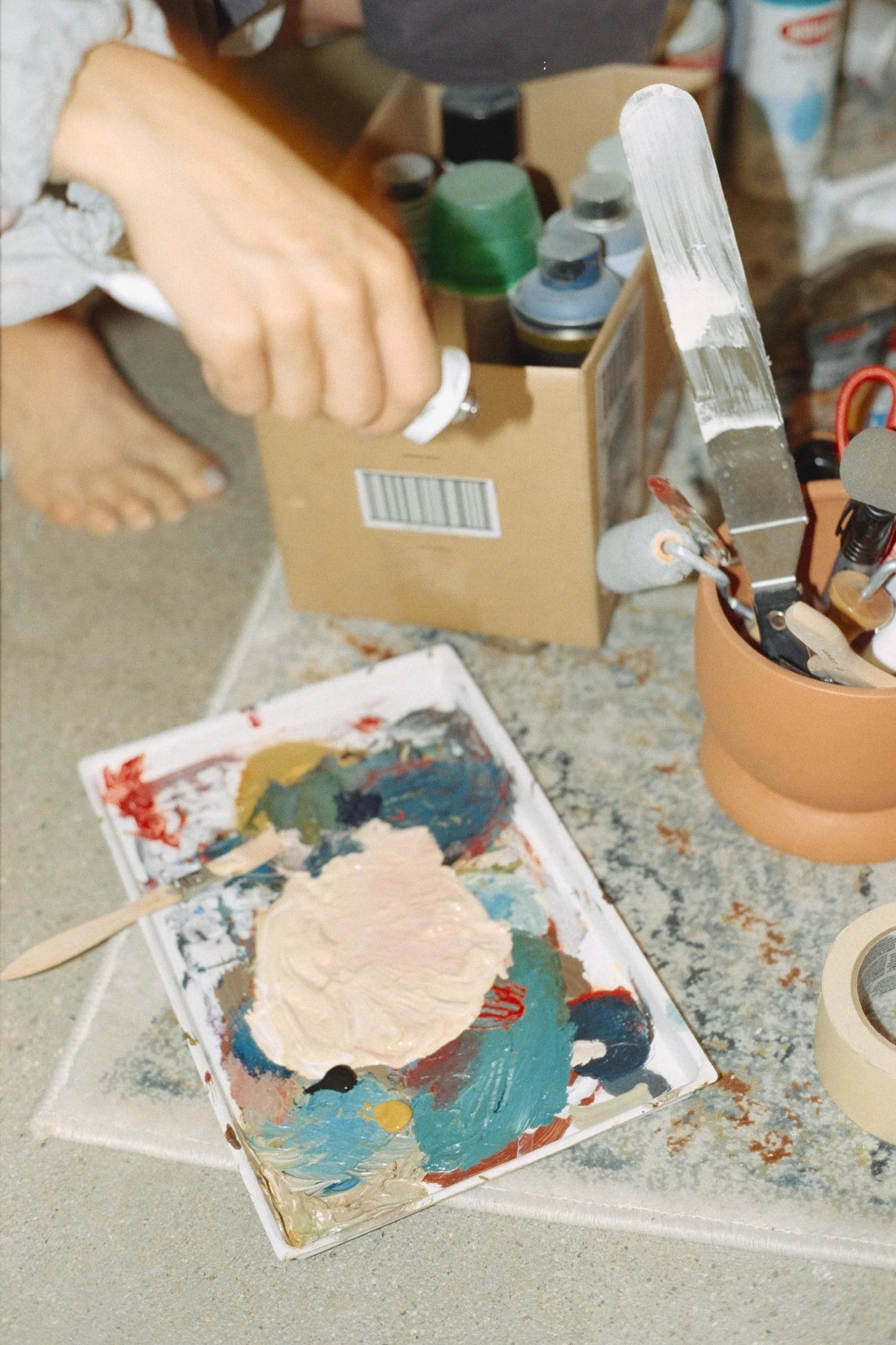 A person's hand holding a small white container, next to a cardboard box filled with spray paint cans, a palette with mixed paints, and a container holding paintbrushes and tools.