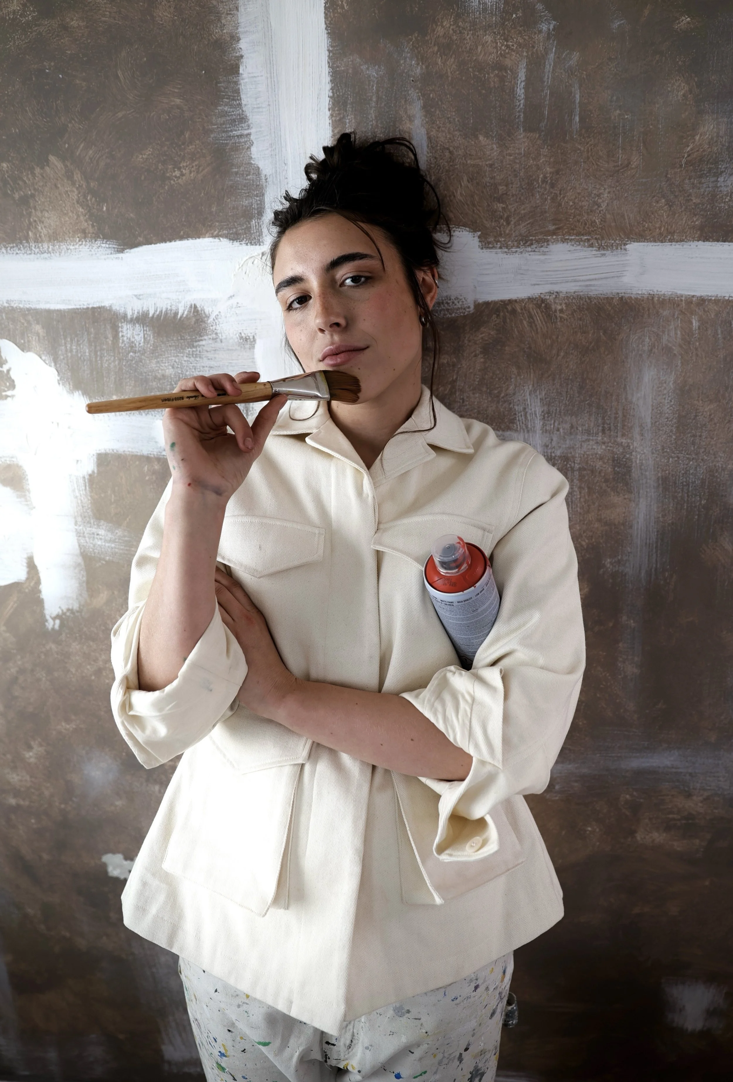 Young woman with dark hair tied up, wearing a beige jacket, holding a paintbrush near her face, standing in front of a partially painted brownish wall with white streaks.