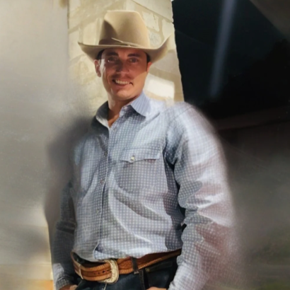 Man wearing a cowboy hat and a light blue plaid shirt, smiling with a light-colored brick wall in the background.