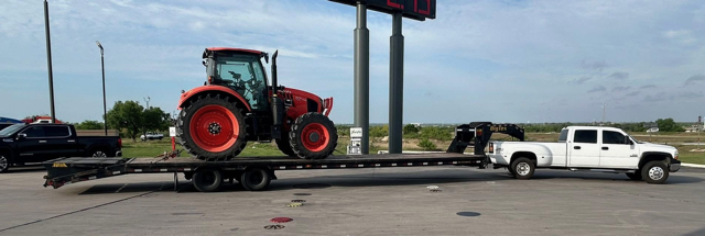 White pickup truck towing a flatbed trailer with a large red tractor on it, parked in a lot under a cloudy sky.