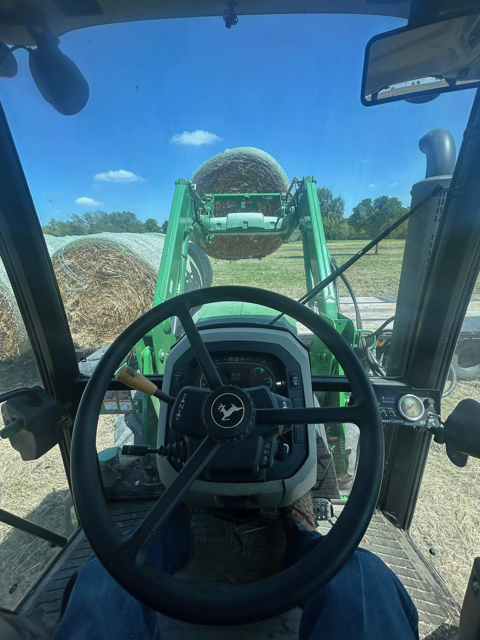 Inside the cab of a tractor, looking through the windshield at a large round hay bale being lifted by a front loader attachment outside.