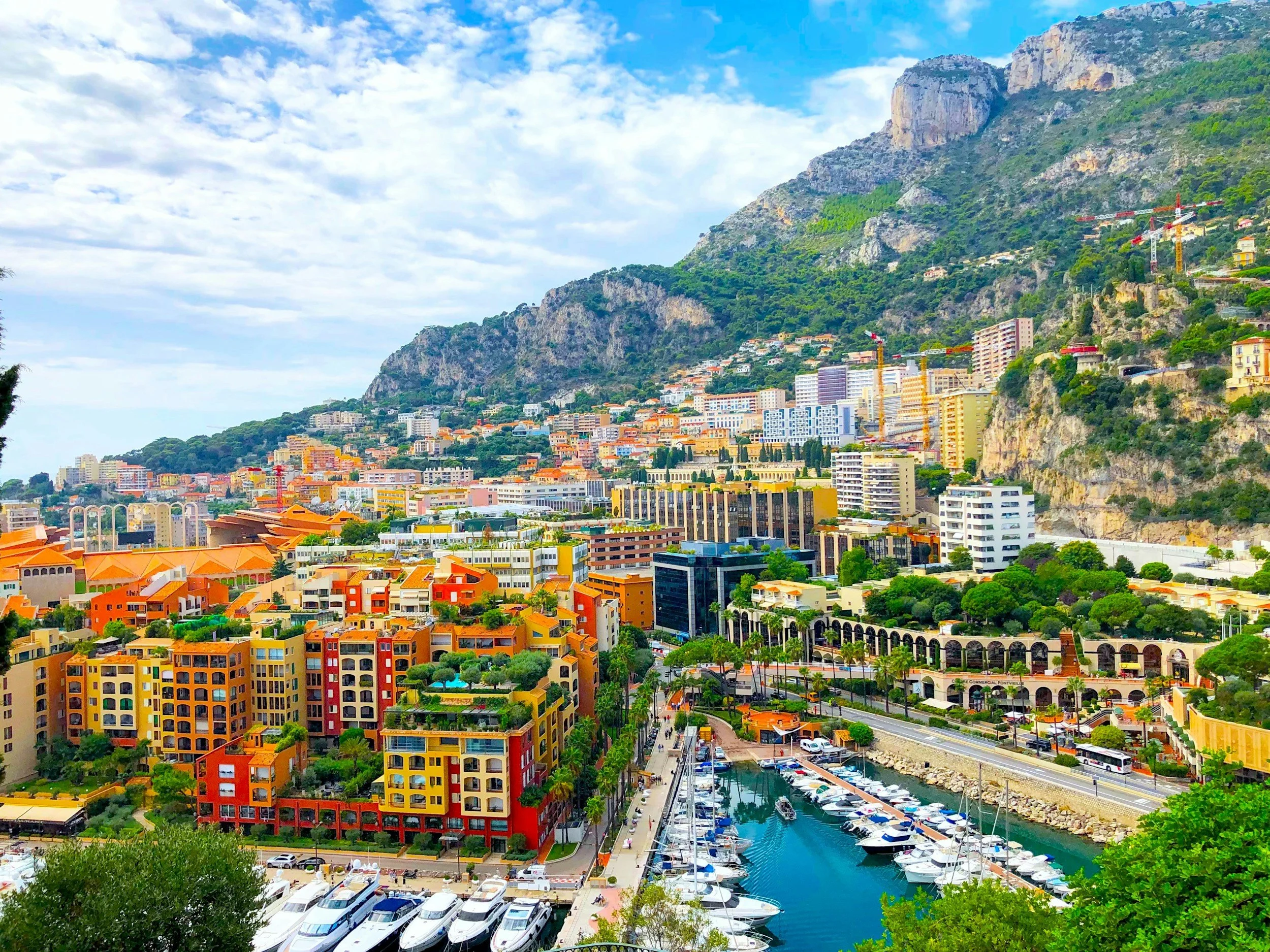 Colorful buildings along a marina with boats, overlooking a mountain hillside with green trees and construction cranes, under a partly cloudy sky.