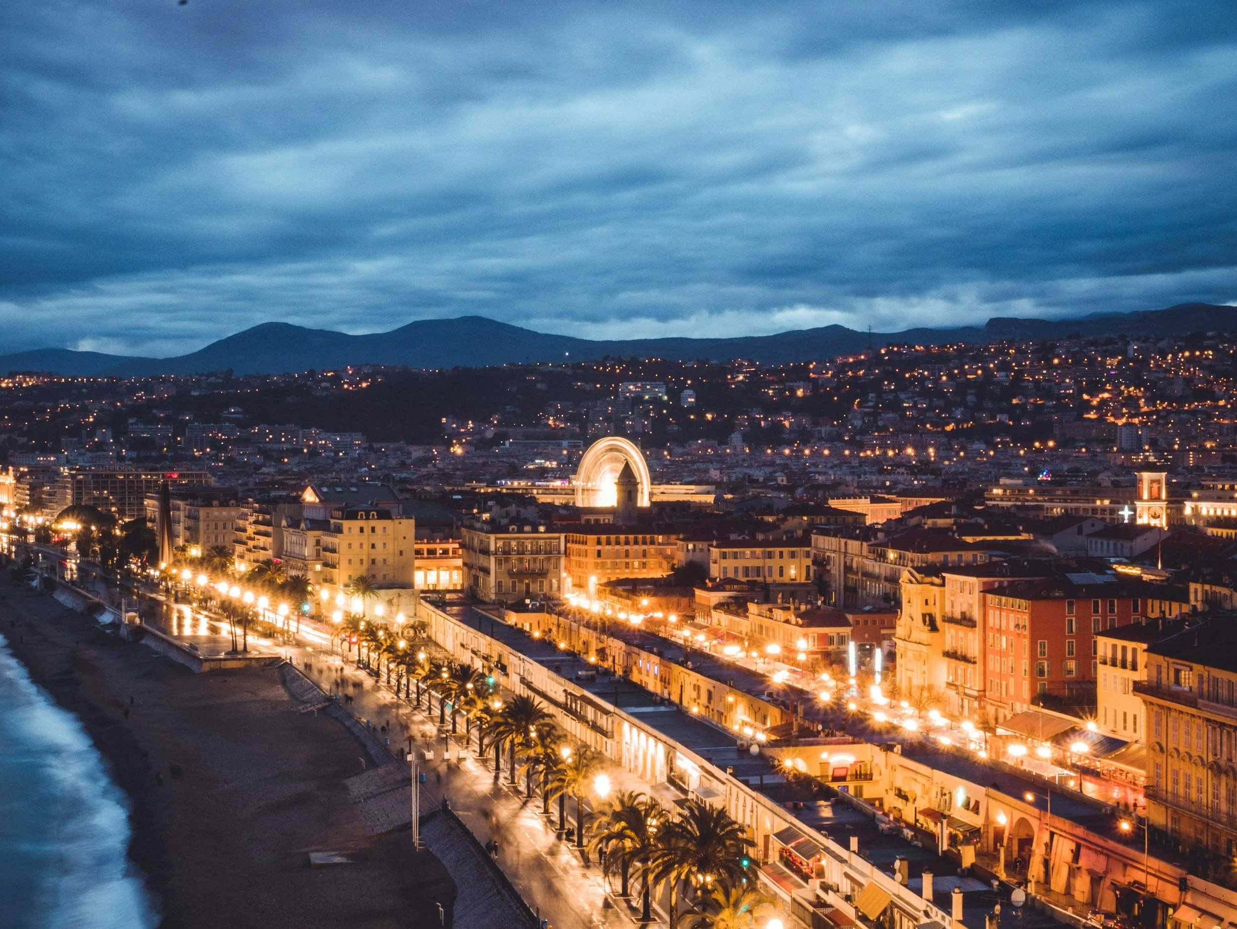 A cityscape at dusk with illuminated buildings along the coast and a mountain range in the background, cloudy sky overhead.
