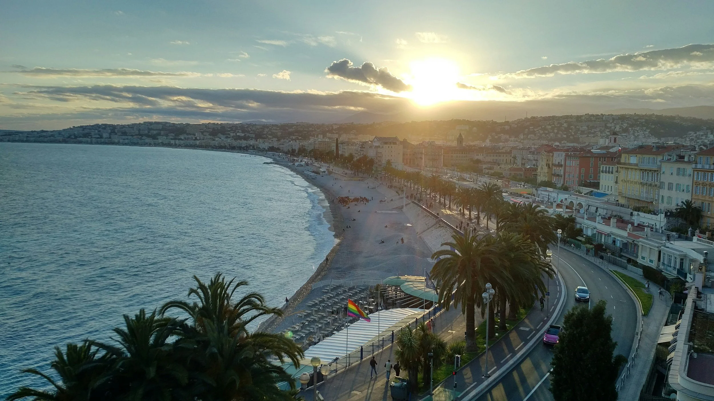 Sunset over a coastal city with colorful buildings, palm trees lining the waterfront promenade, and a beach with people near the shoreline.