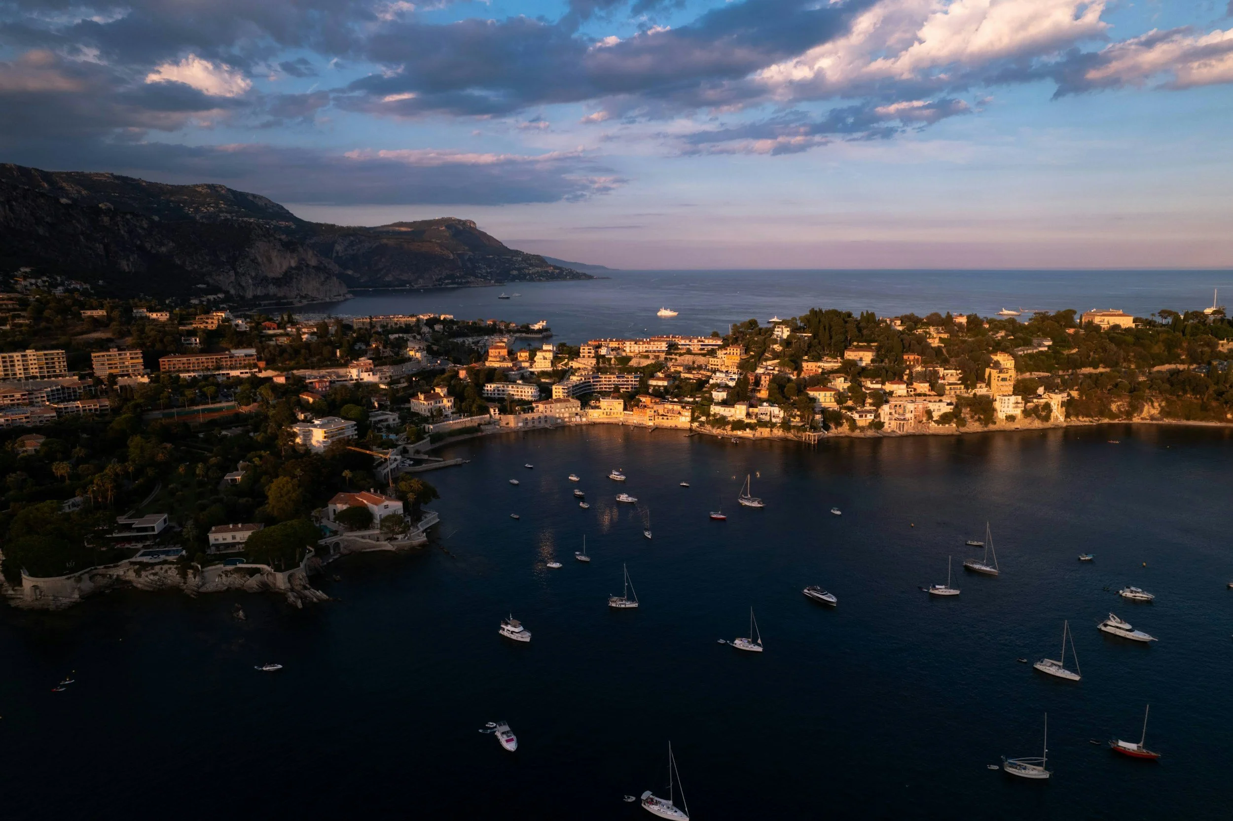 An aerial view of a coastal city with a harbor filled with sailboats and yachts, high-rise buildings along the shoreline, rolling hills behind the city, and a mountainous coastline in the distance under a partly cloudy sky.