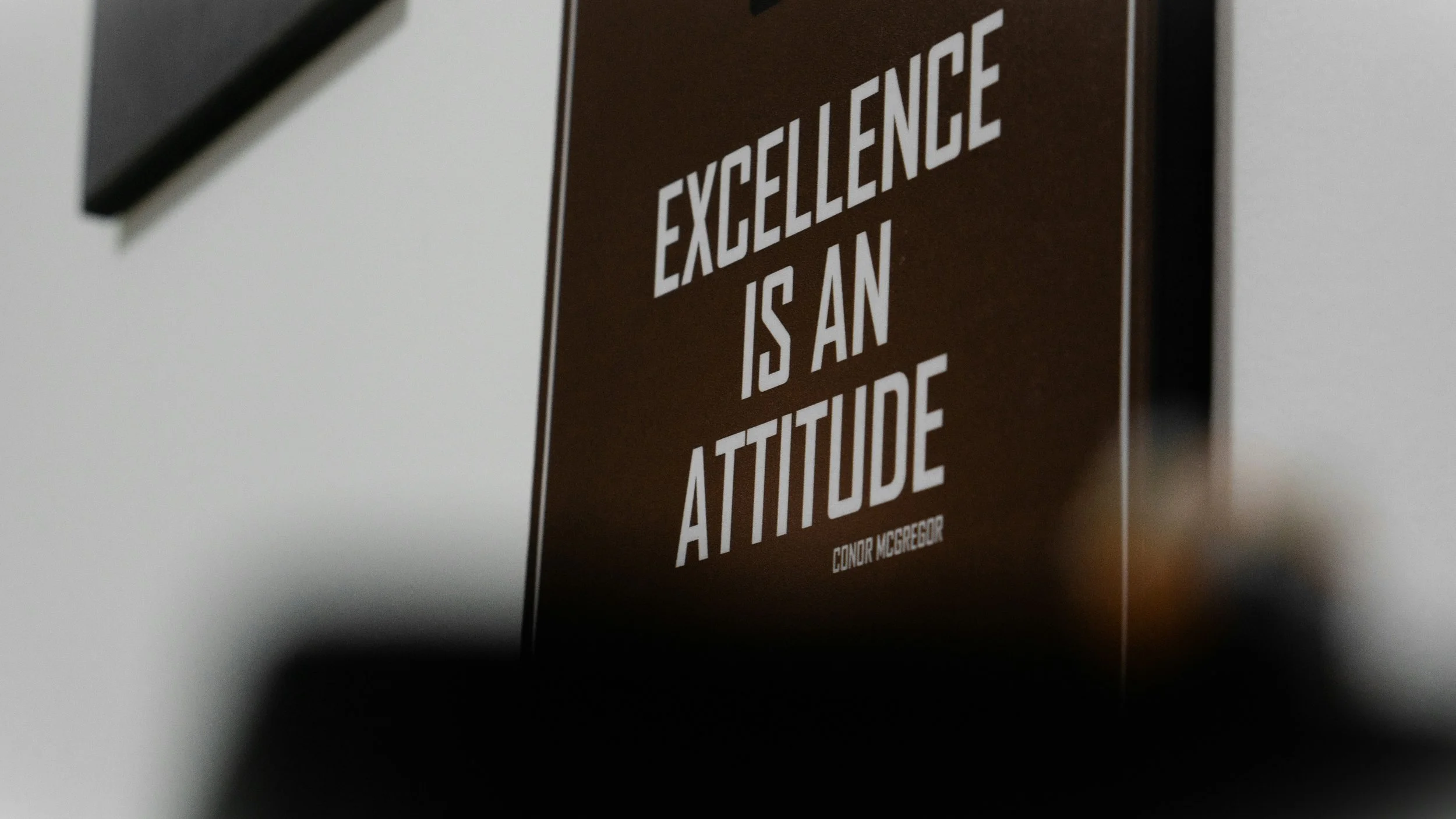 Close-up of a brown poster on a wall with the quote 'Excellence is an attitude' by Connor McGregor in white bold uppercase letters.