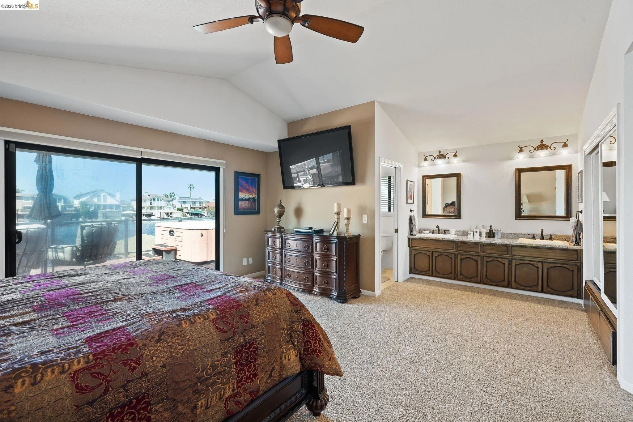 Bedroom with sliding glass door leading to balcony, bed with patterned quilt, dresser with decorative items, wall-mounted TV, double sink vanity with mirrors and lighting, and small bathroom area.