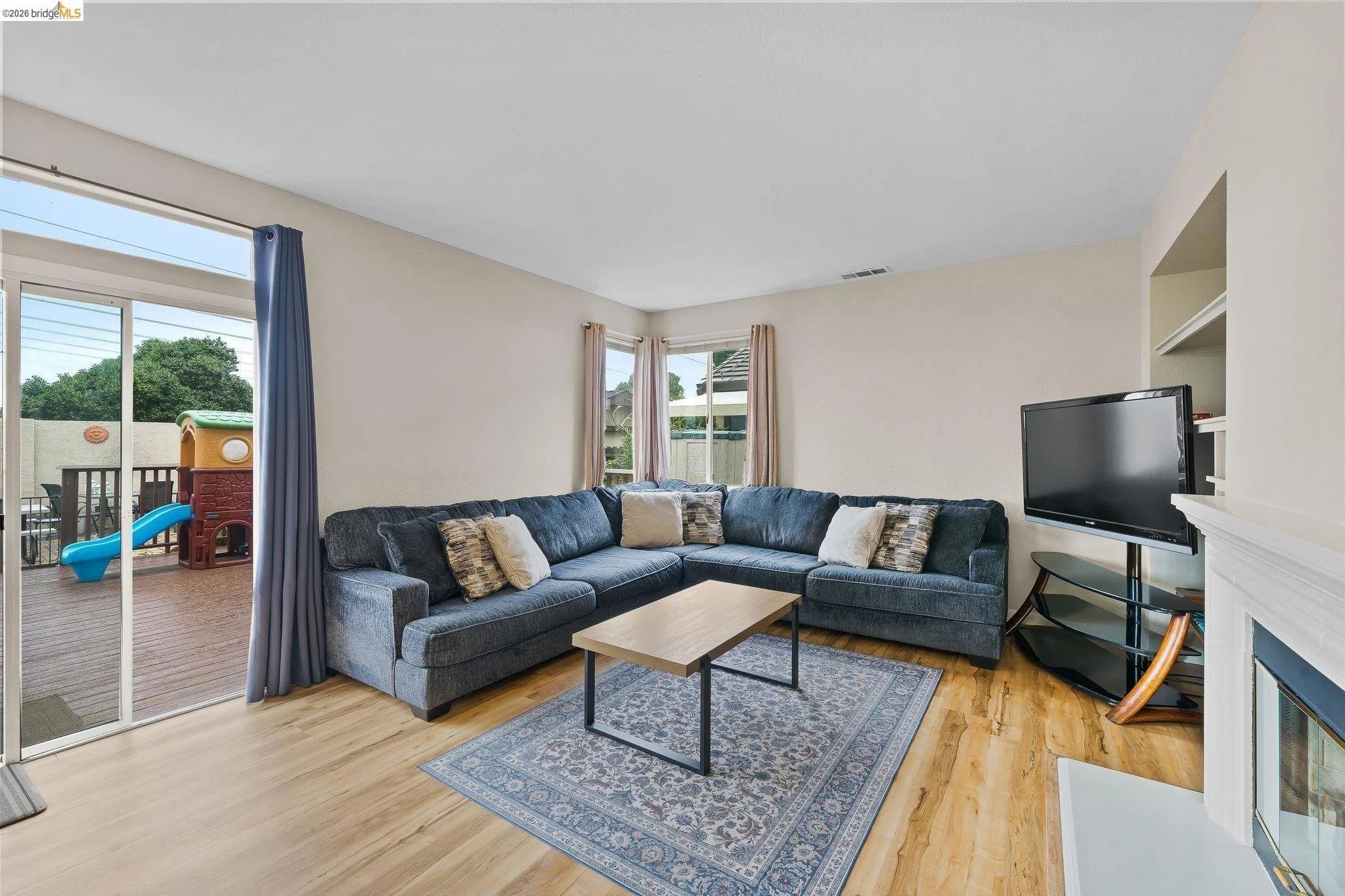 Living room with a large L-shaped gray sofa, a wooden coffee table on a patterned rug, a flat-screen TV on a black stand, windows with beige curtains, and a sliding glass door leading to a patio with outdoor play equipment.