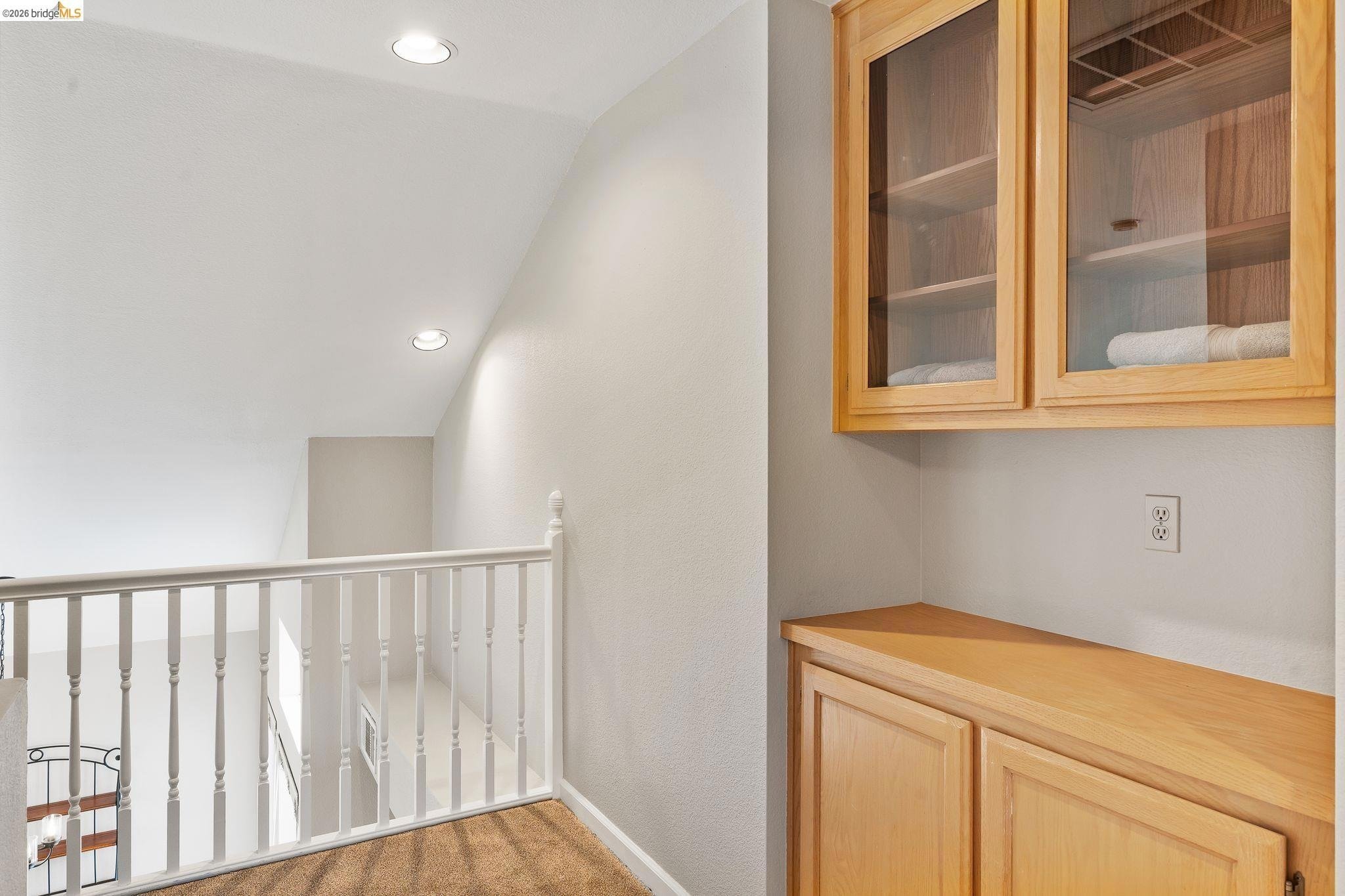 Wooden cabinet with glass doors and towels inside, against white wall near staircase railing in a well-lit hallway.