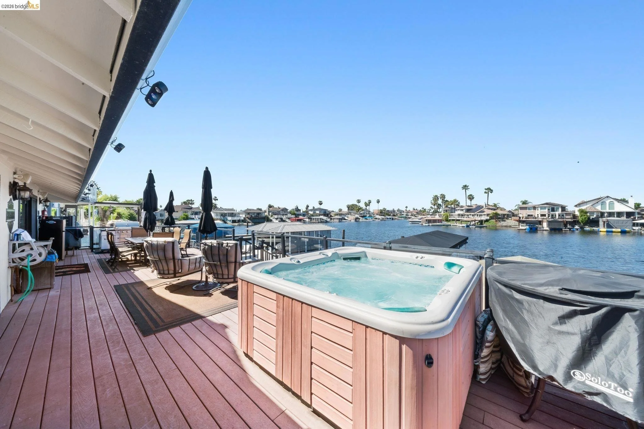 Deck with hot tub, outdoor dining table, chairs, and umbrellas overlooking a waterway with boats and houses in the distance.