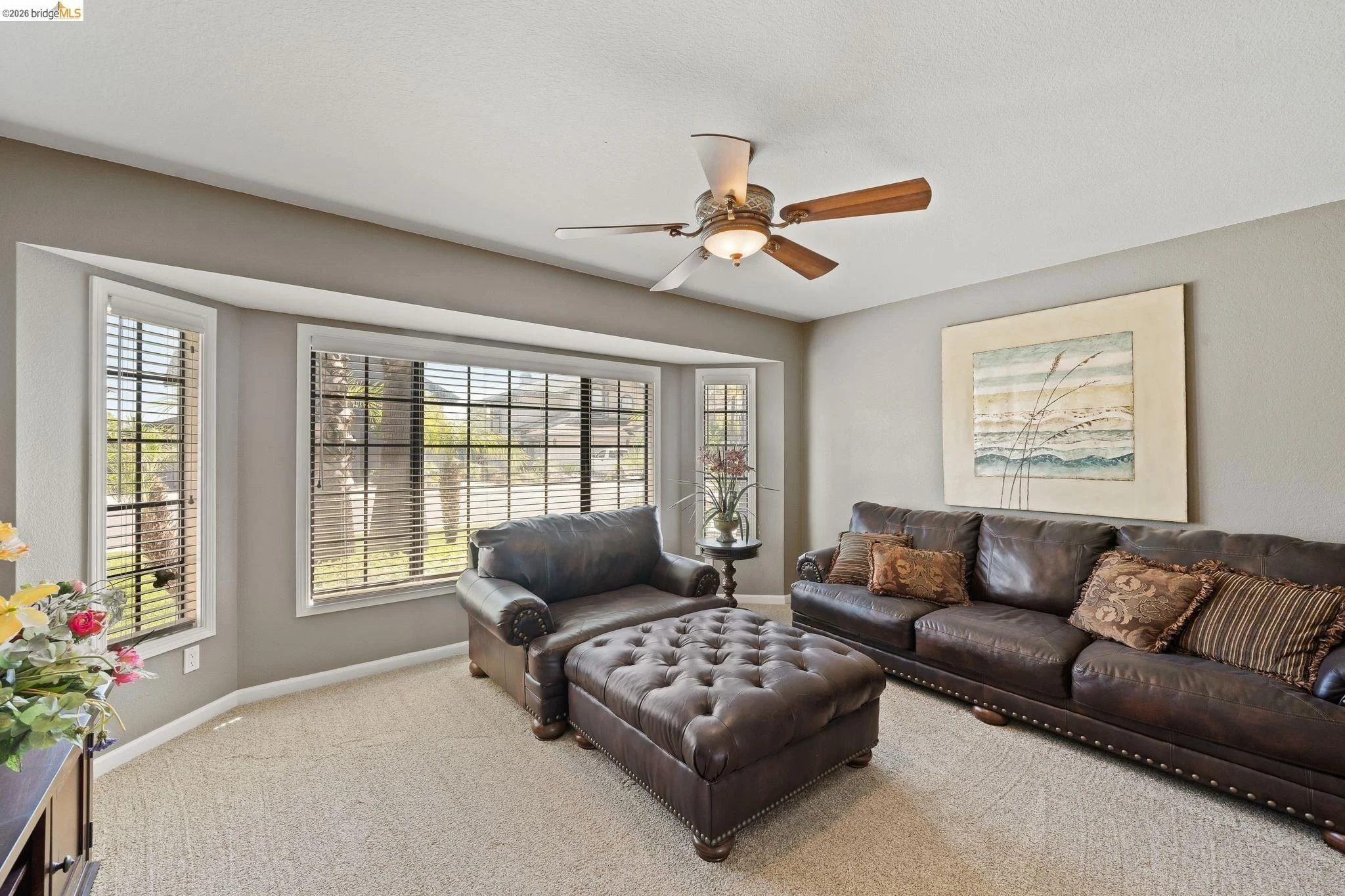 Living room with large bay window, leather sofa, matching armchair, tufted ottoman, ceiling fan, and artwork on the wall.