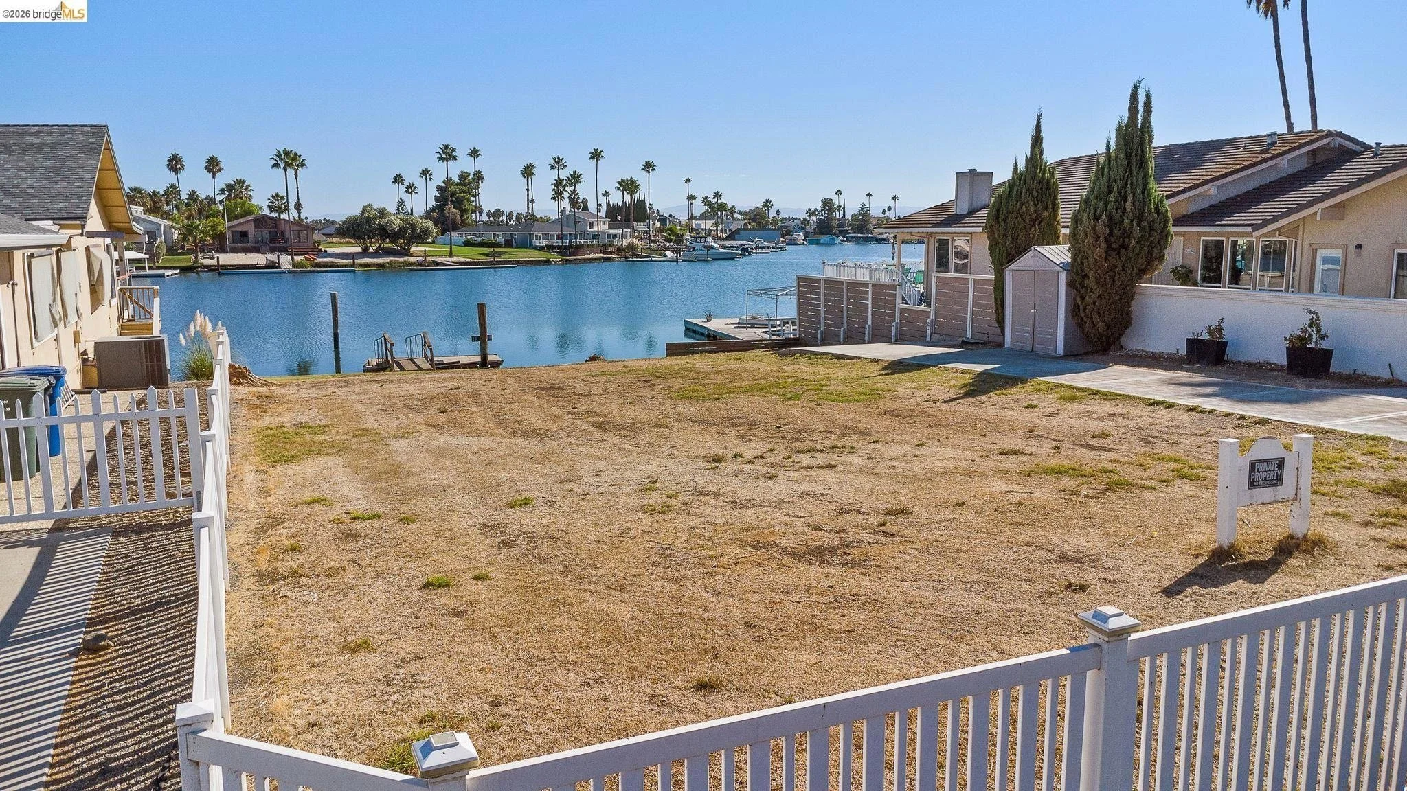 A fenced backyard with a view of a waterway, houses, boats, and palm trees in the background, under a blue sky.