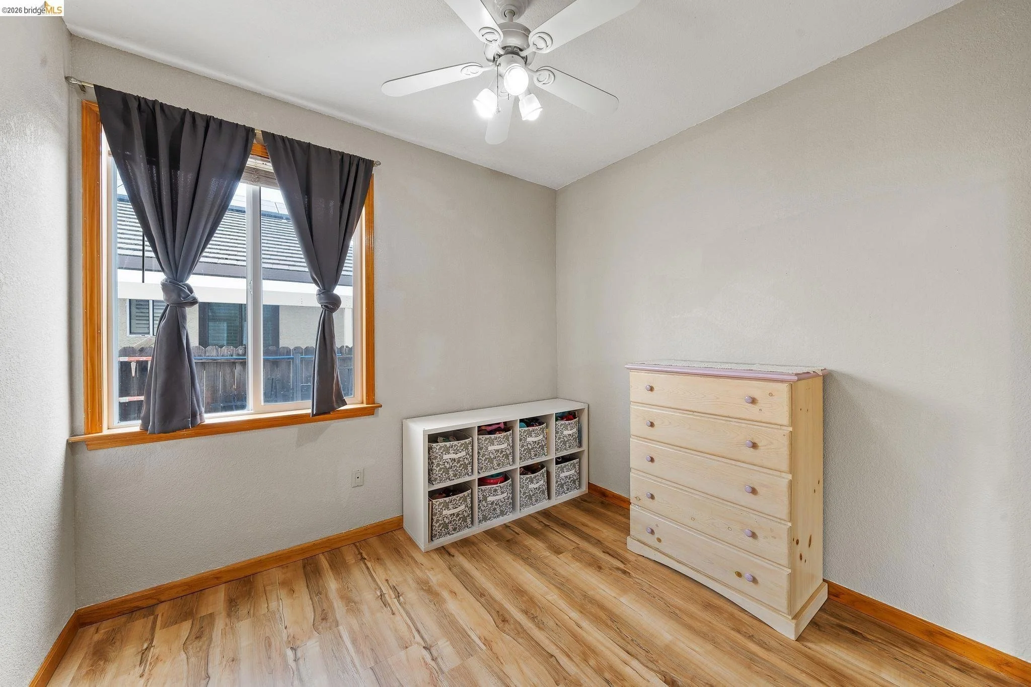 Empty bedroom with hardwood floors, a window with dark curtains, a small white storage cube, and a light-colored dresser.