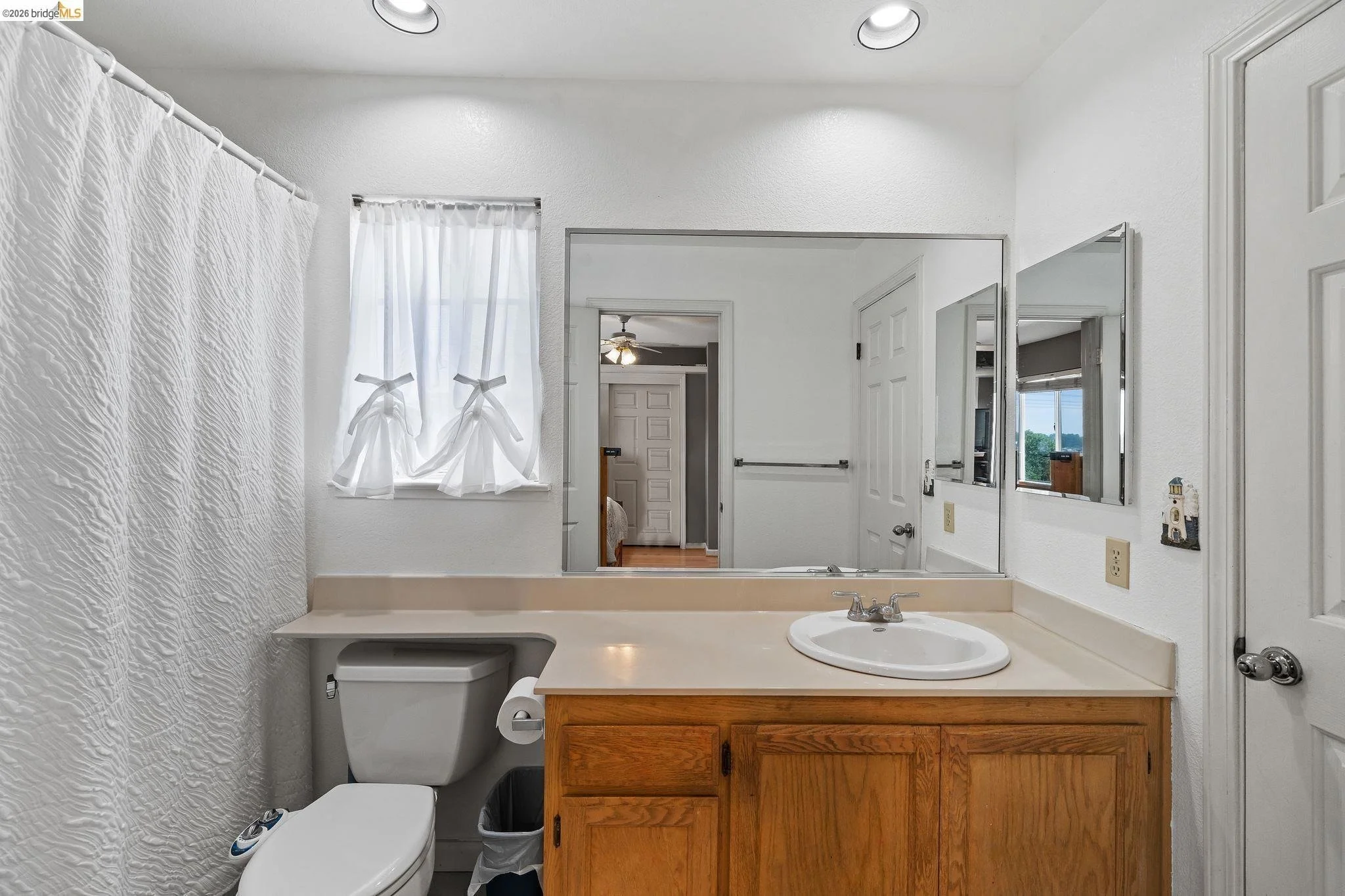 Bathroom with a toilet, a sink, and a large mirror, white textured shower curtain, window with white curtains, and a door leading to another room.