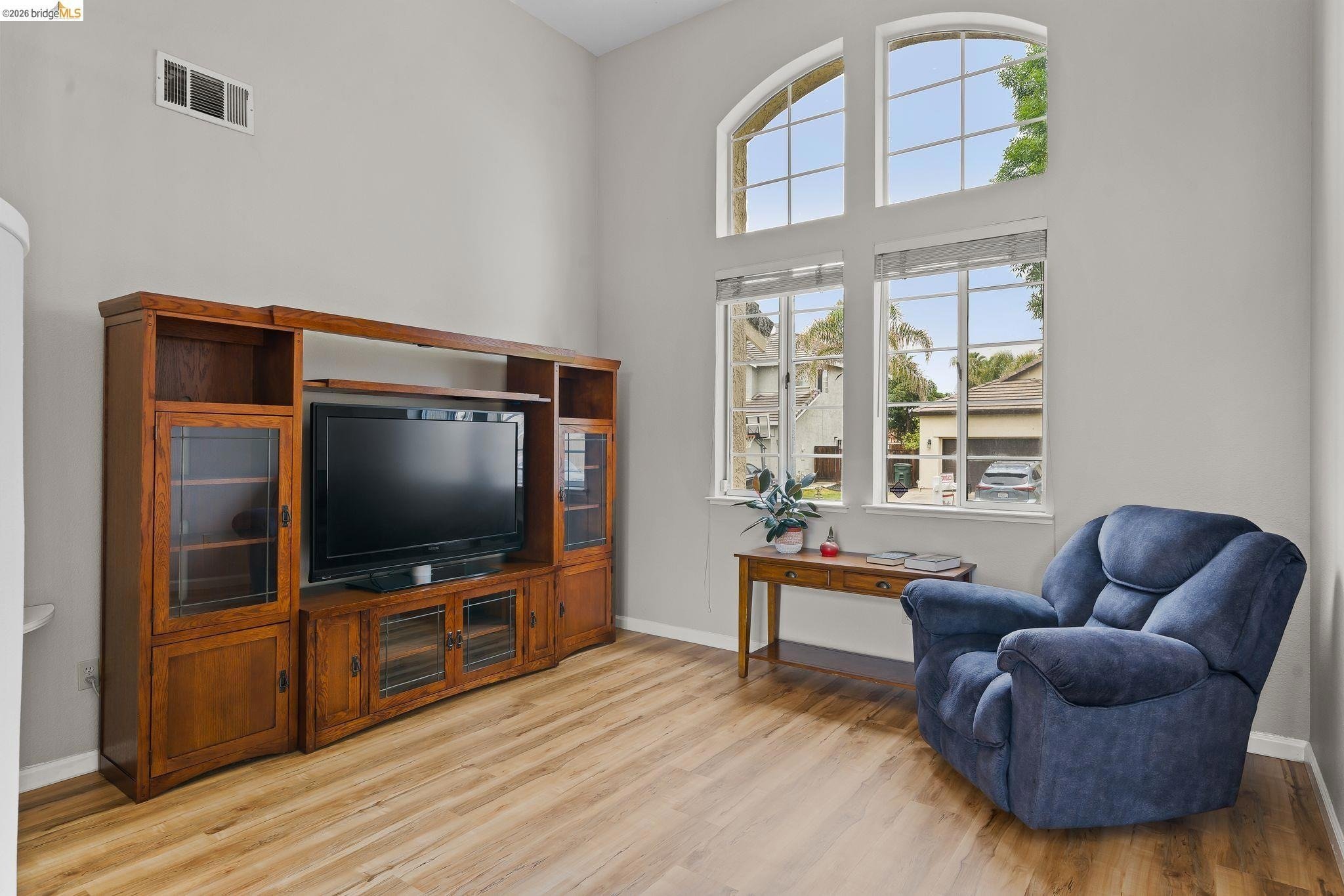 Living room with large arched windows, a blue armchair, a wooden entertainment center with a TV, and a small wooden side table with books and a potted plant.