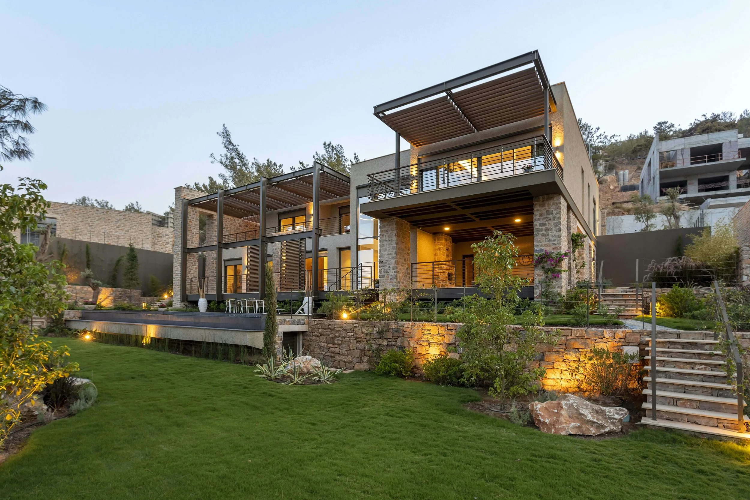 Modern multi-story house with stone and concrete exterior, illuminated in soft evening light, surrounded by a landscaped yard with grass, plants, and stone pathways.