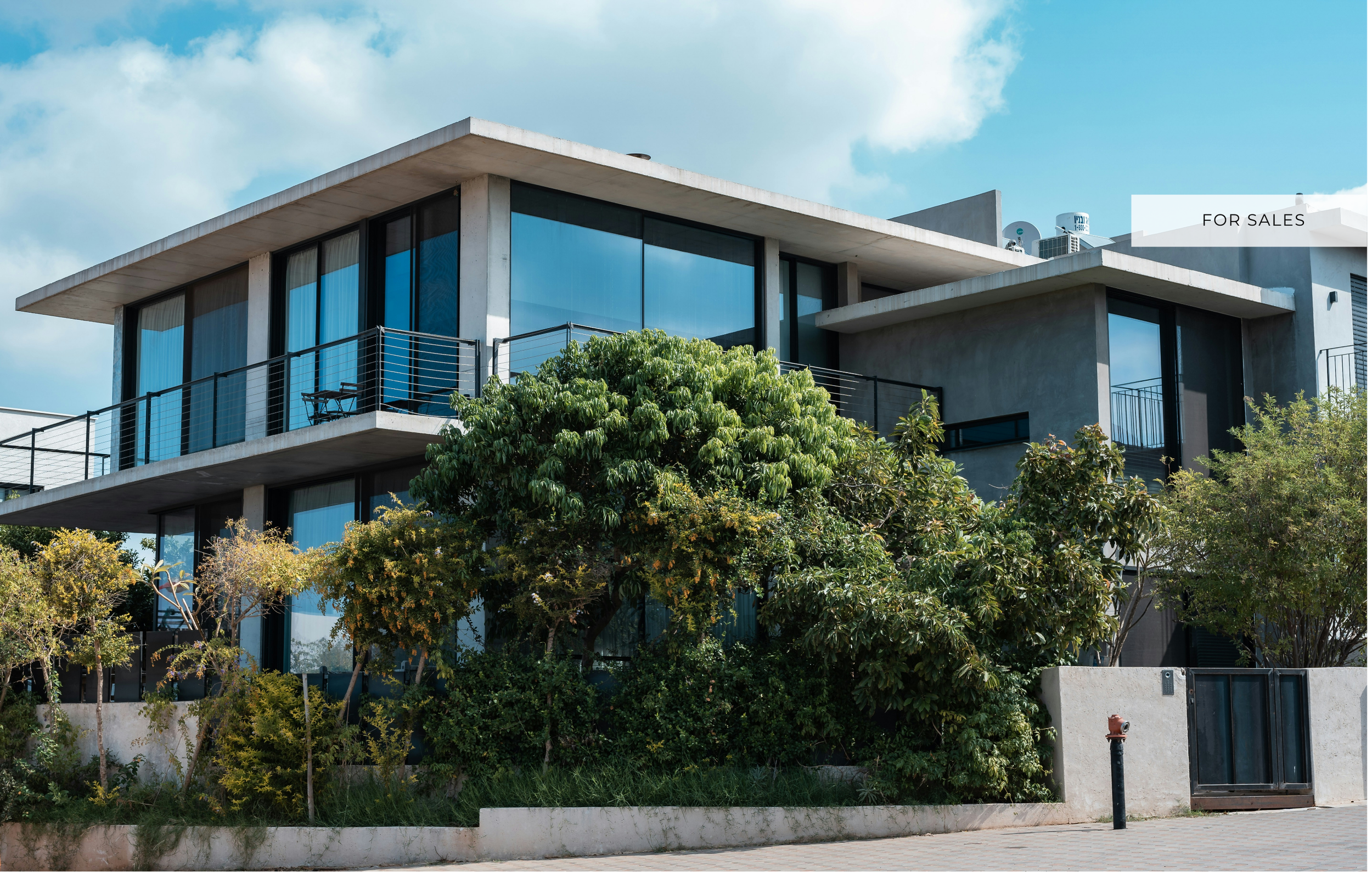 Modern multi-story house with large glass windows, balconies, surrounded by greenery and trees, with a partly cloudy sky in the background.