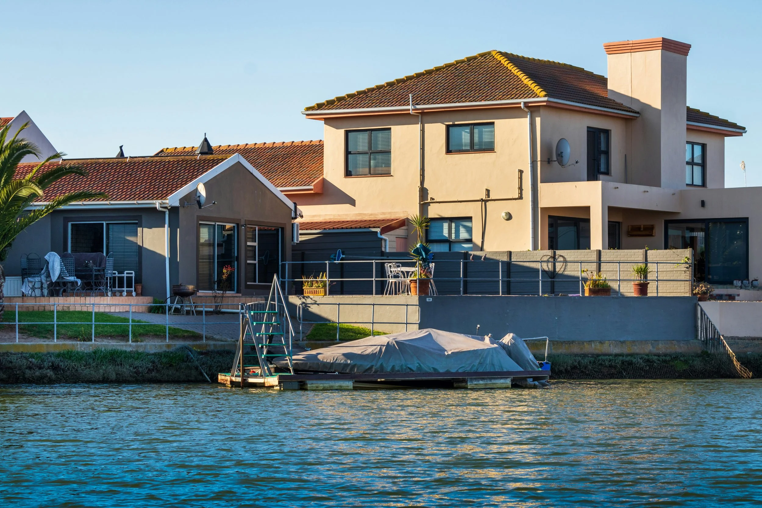 Residential house by a waterway with boats and boat dock, outdoor furniture, and potted plants.