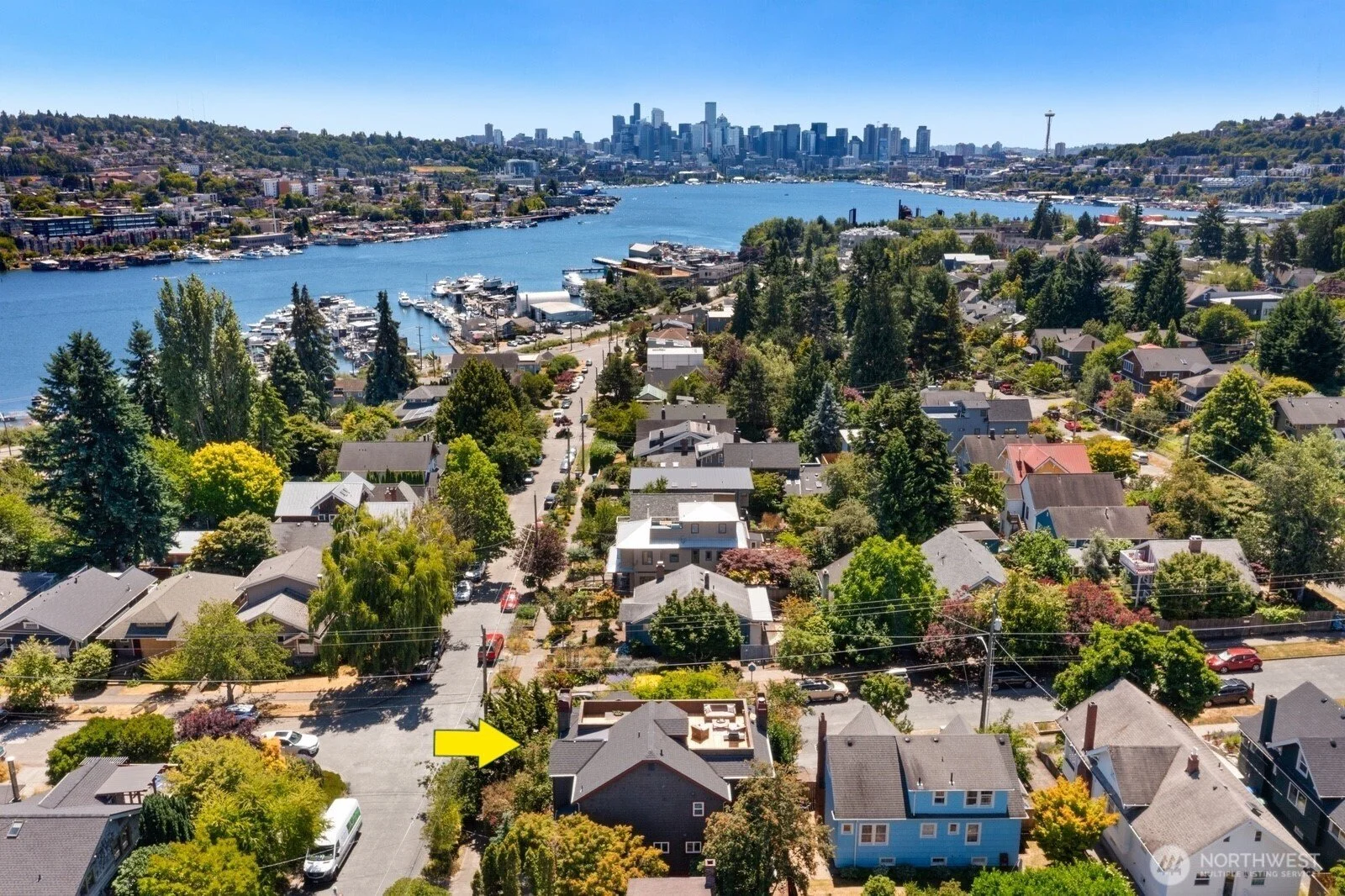 Aerial view of a neighborhood near a body of water with boats, houses, and trees, overlooking a city skyline in the background.