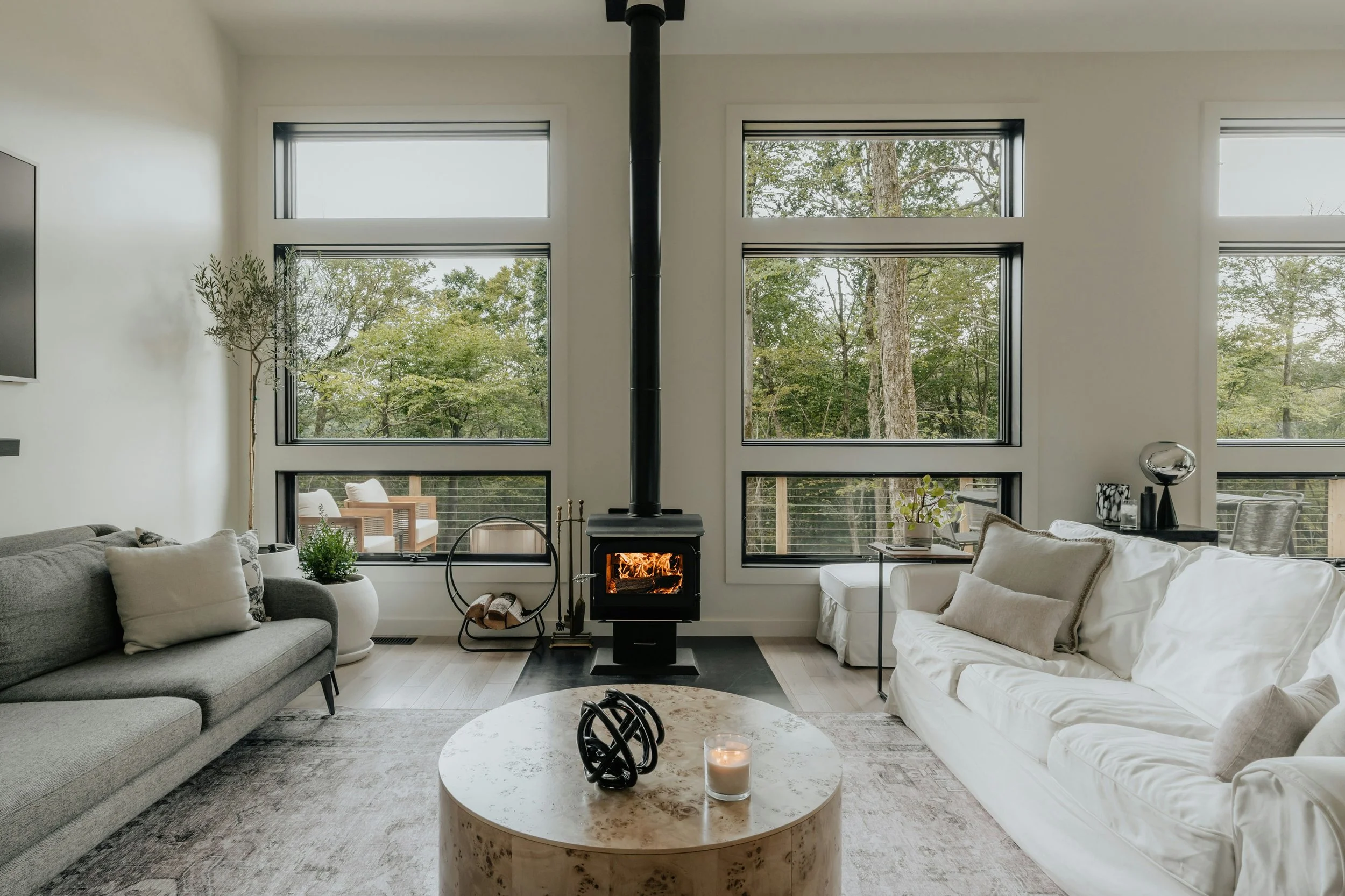 Modern living room with gray and white sofas, a black wood stove emitting flames, large windows showing trees outside, a round marble coffee table with decorative black sculpture and candle, and various indoor plants and decor.