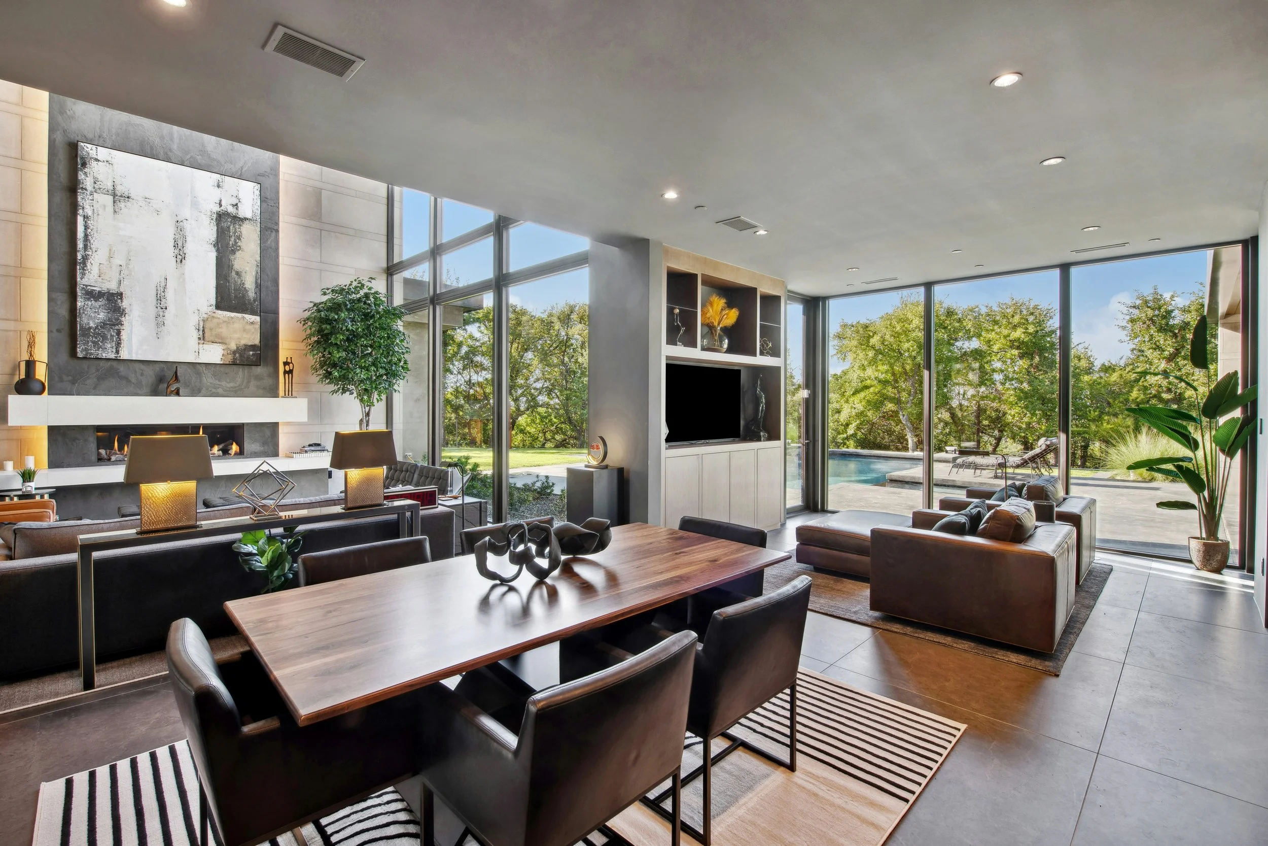 Modern living room with large floor-to-ceiling windows, a wooden dining table with black chairs, a sitting area with leather sofa and ottoman, a fireplace, abstract artwork, potted plants, and a view of the backyard with trees, a pool, and outdoor se