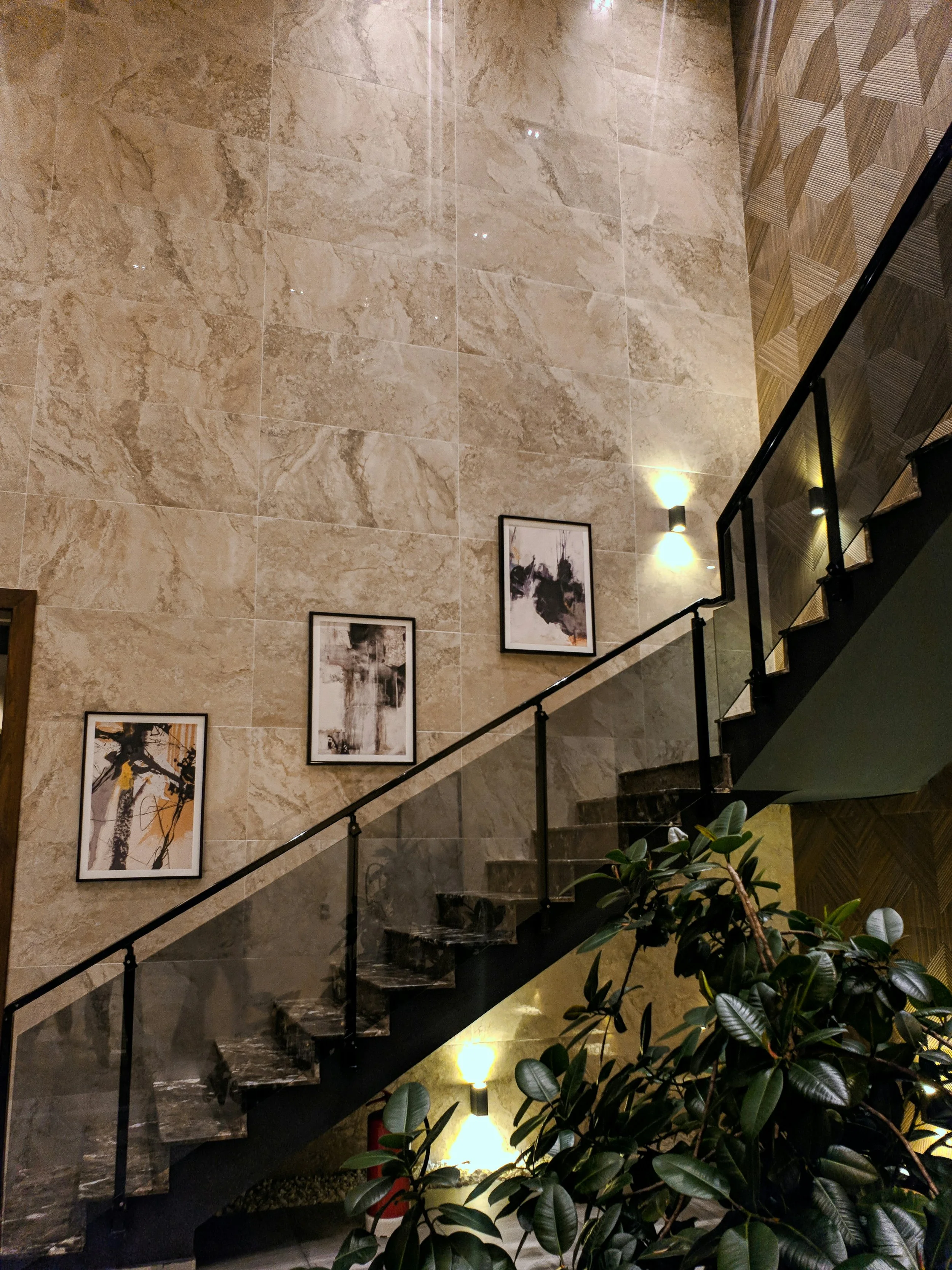 Interior view of a staircase with a black railing and marble steps, against a beige marble wall decorated with three framed abstract art prints. Green leafy plant in the foreground, warm wall lights illuminate the area.