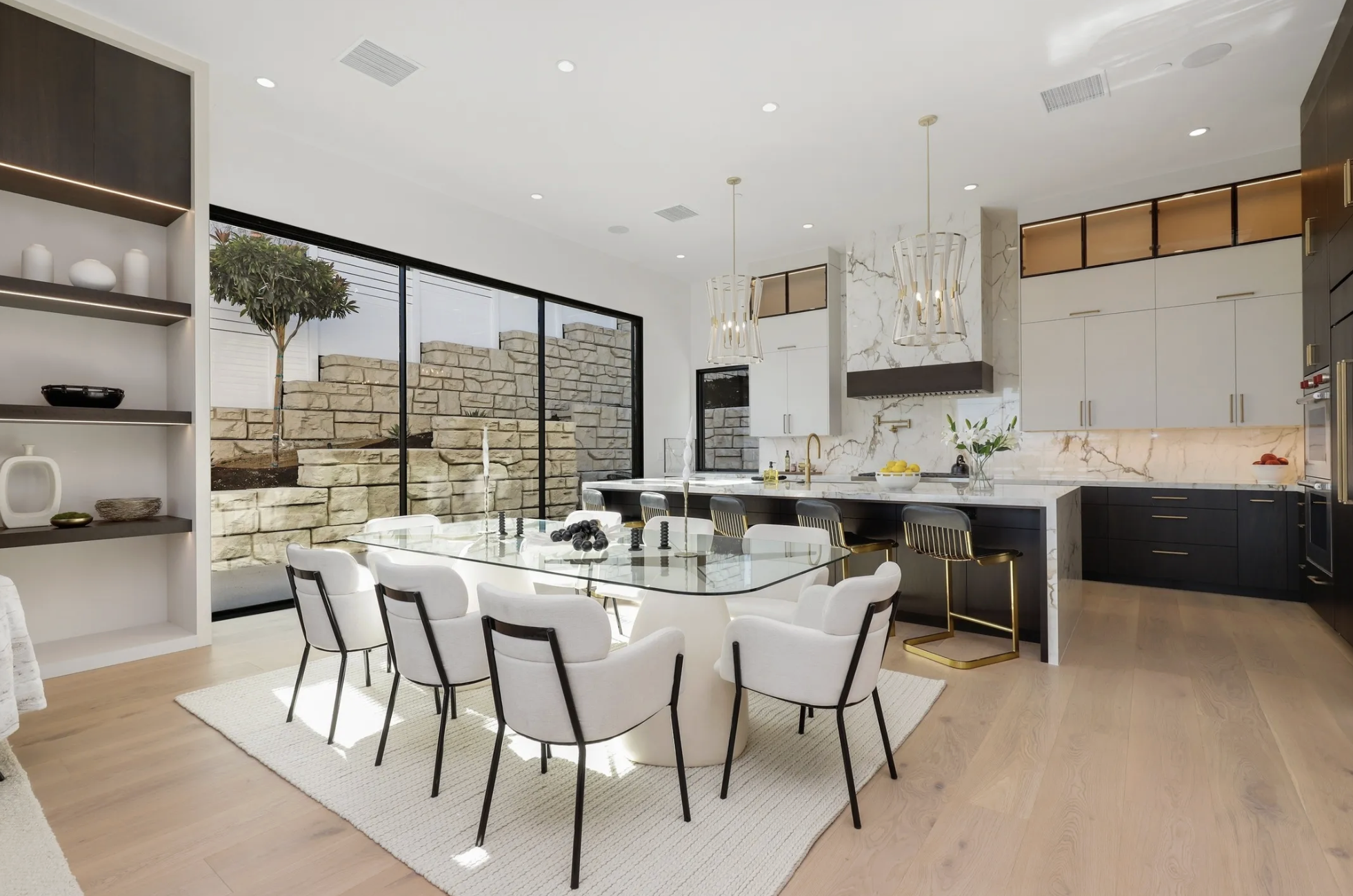 Modern open-concept kitchen and dining area with a glass dining table, white chairs, black and gold barstools, marble countertops, and large sliding glass doors with a view of a stone retaining wall outside.