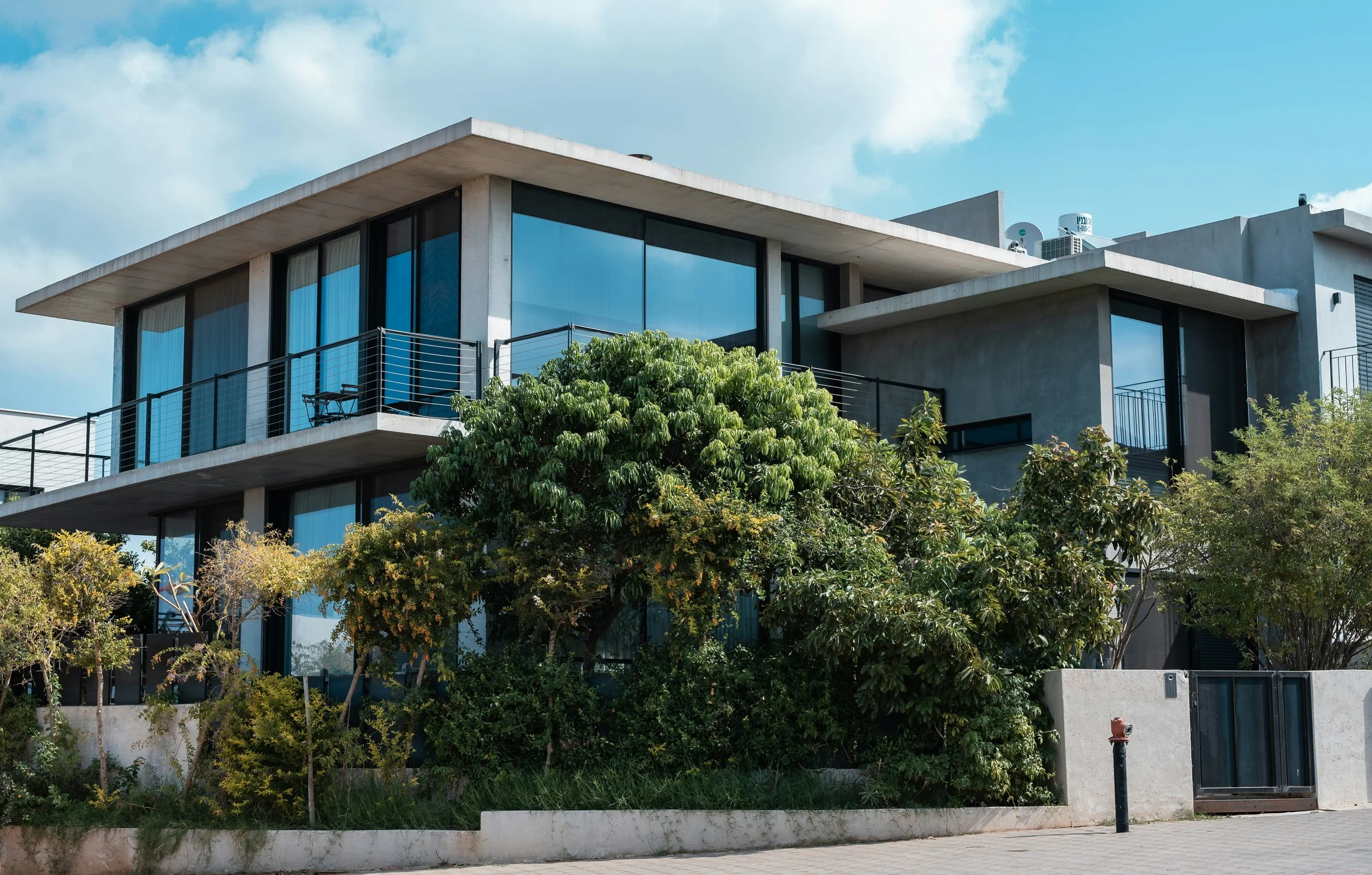 Modern concrete house with large glass windows and balconies, surrounded by green trees and shrubs, on a clear day with a blue sky and some clouds.