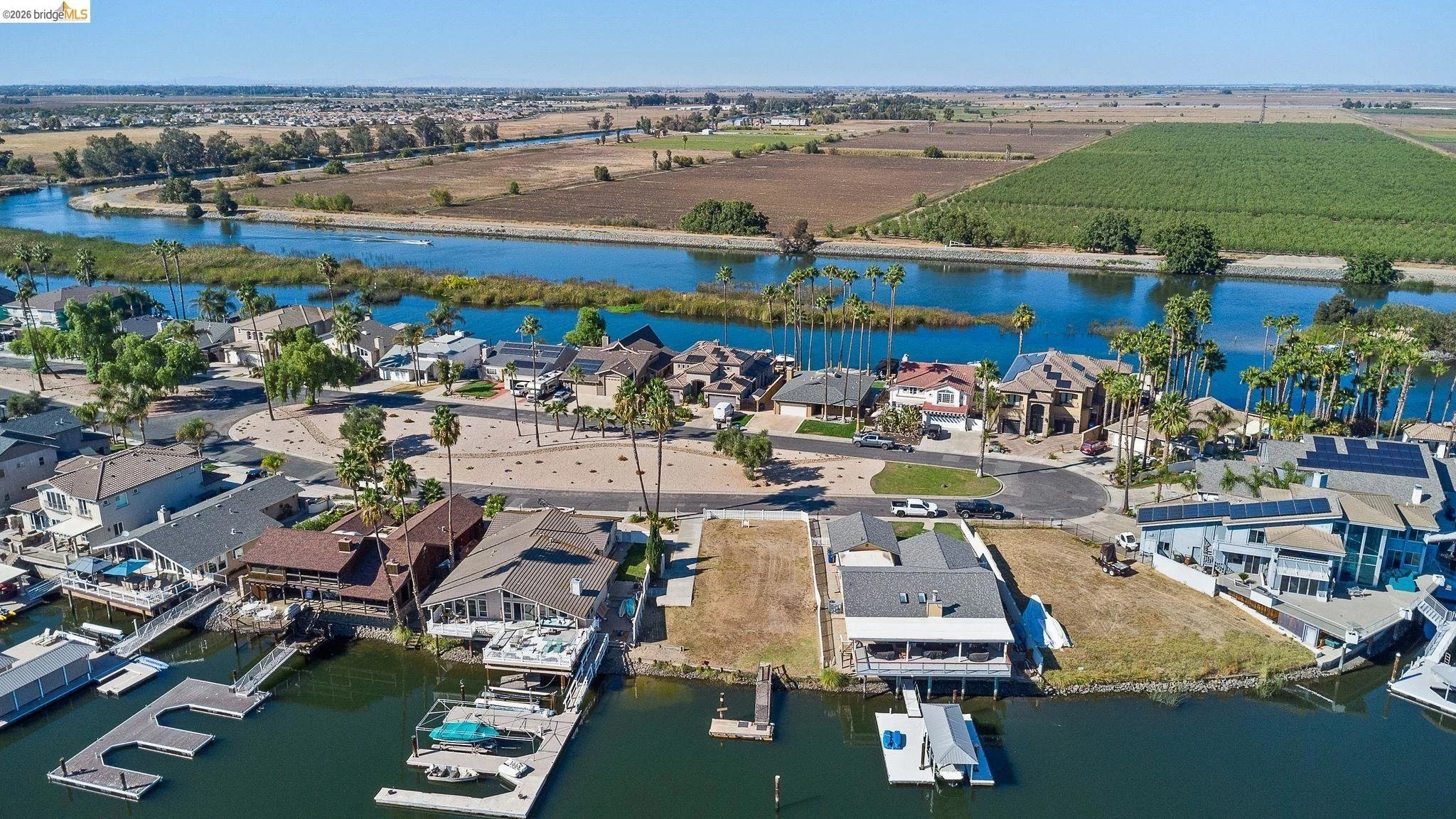 Aerial view of waterfront houses along a canal with boat docks, a residential street, a river with green islands, and farmland in the distance.