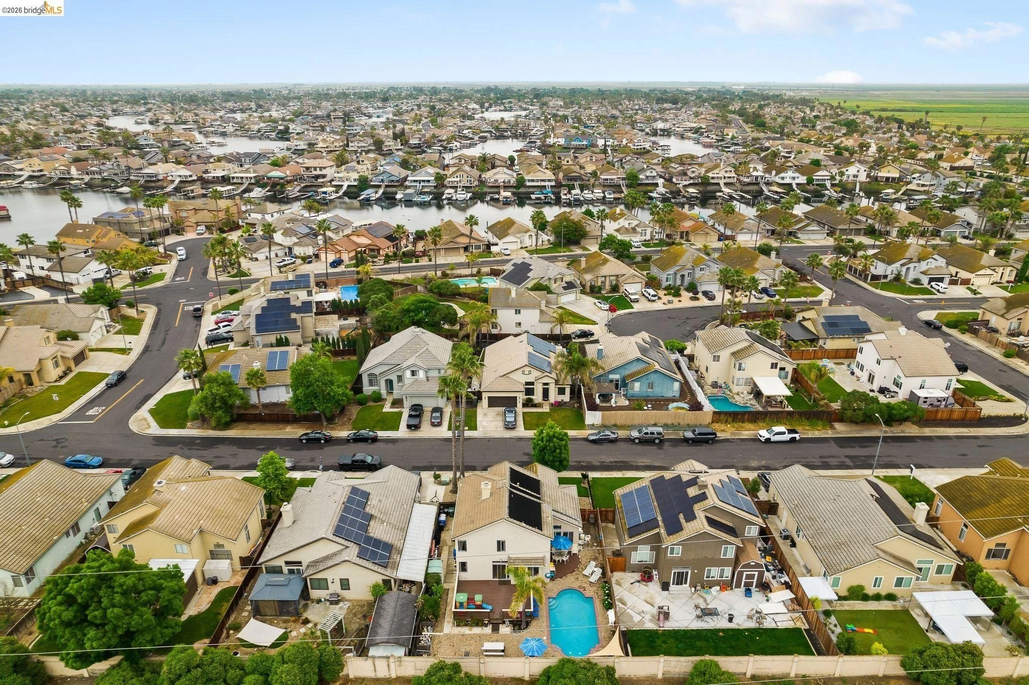 Aerial view of a residential neighborhood featuring houses with solar panels, swimming pools, and streets lined with parked cars. A waterway with boats is visible in the background, alongside open fields and a cloudy sky.