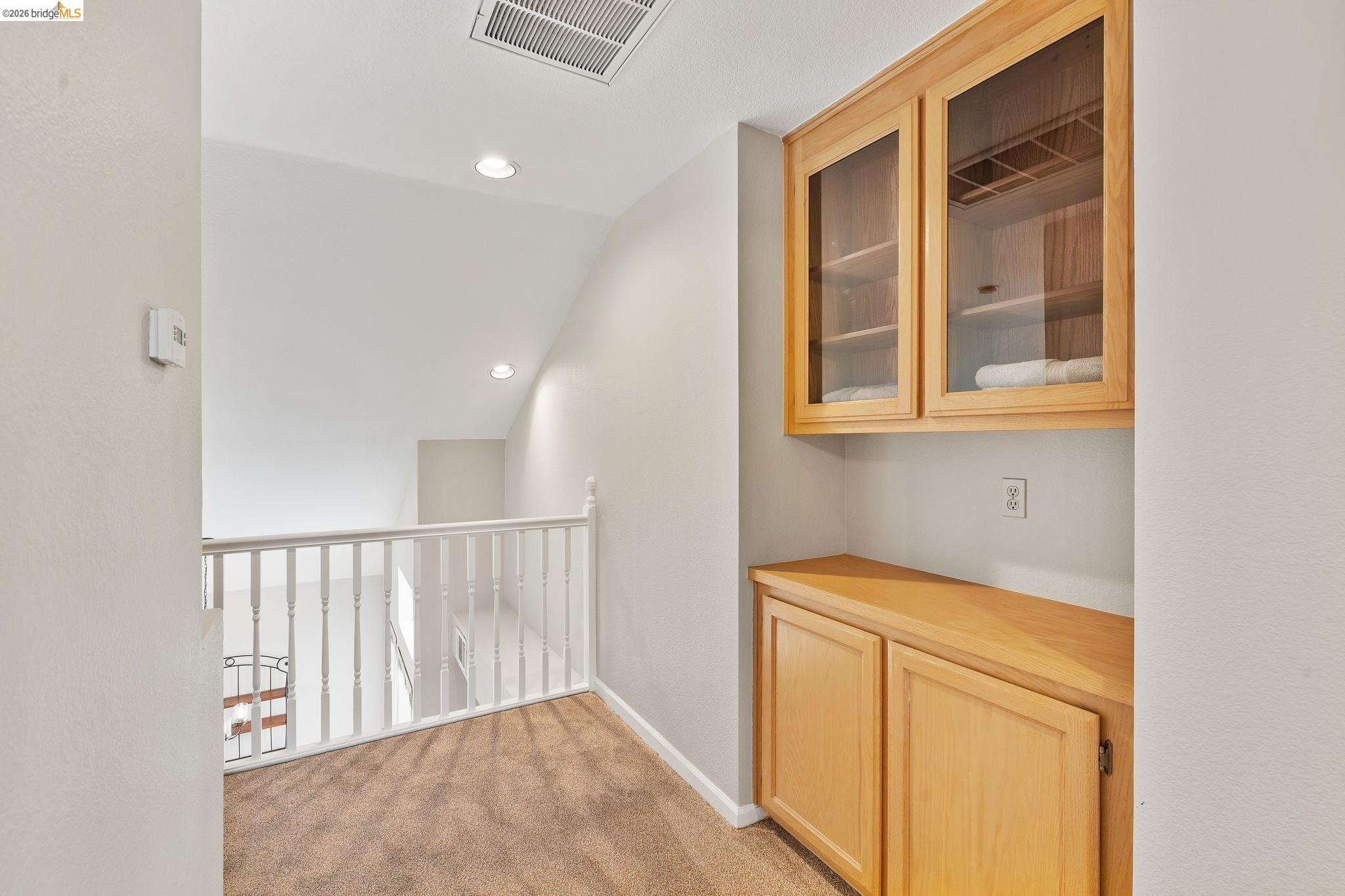 Interior view of a room corner with a wooden cabinet with glass doors and towels, beige carpet, white walls, ceiling vents, and recessed lighting.