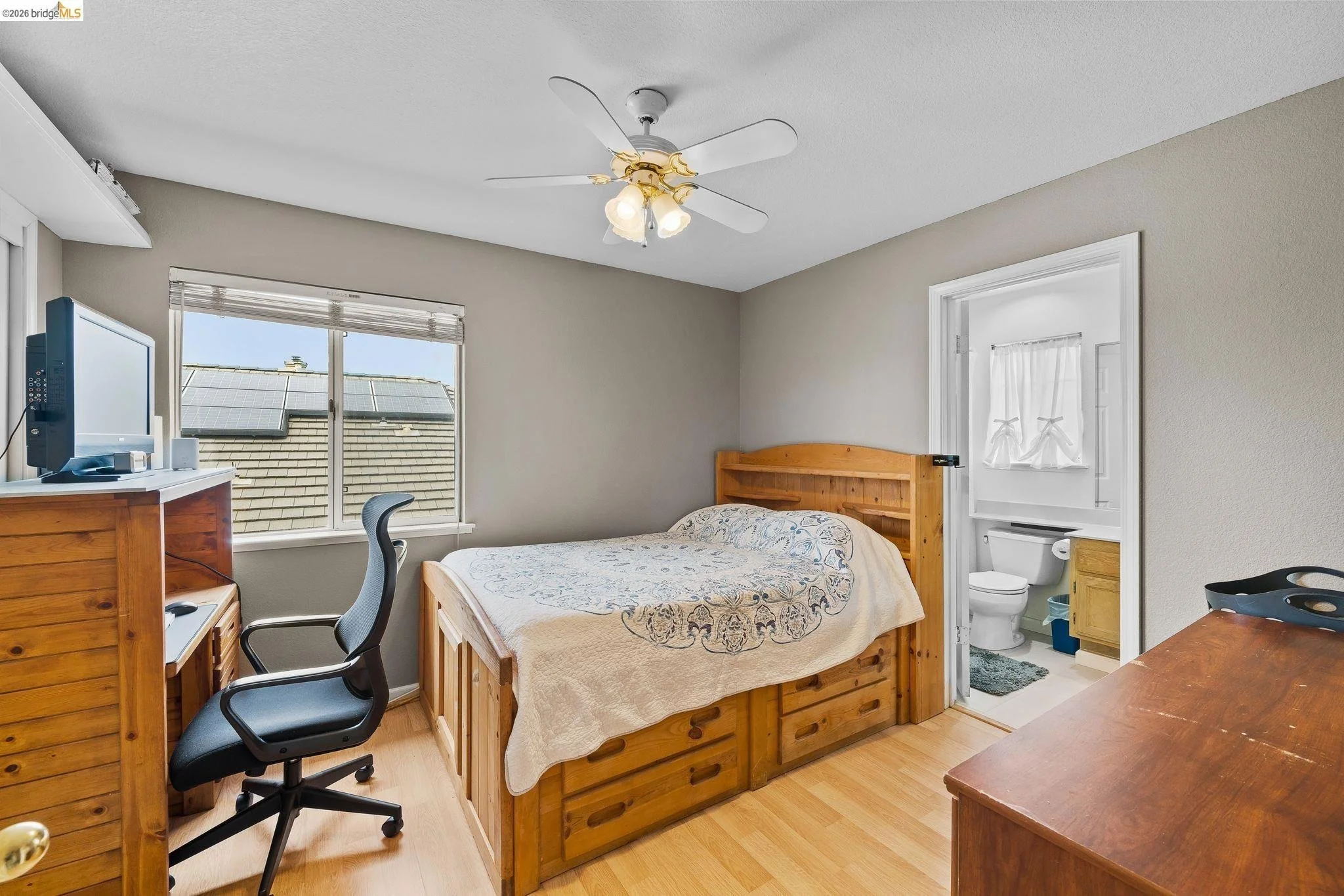Bedroom with a wooden bed, a window showing solar panels on the roof outside, a desk with a computer and a TV, a swivel chair, and an open door leading to a bathroom with a toilet and window with curtains.