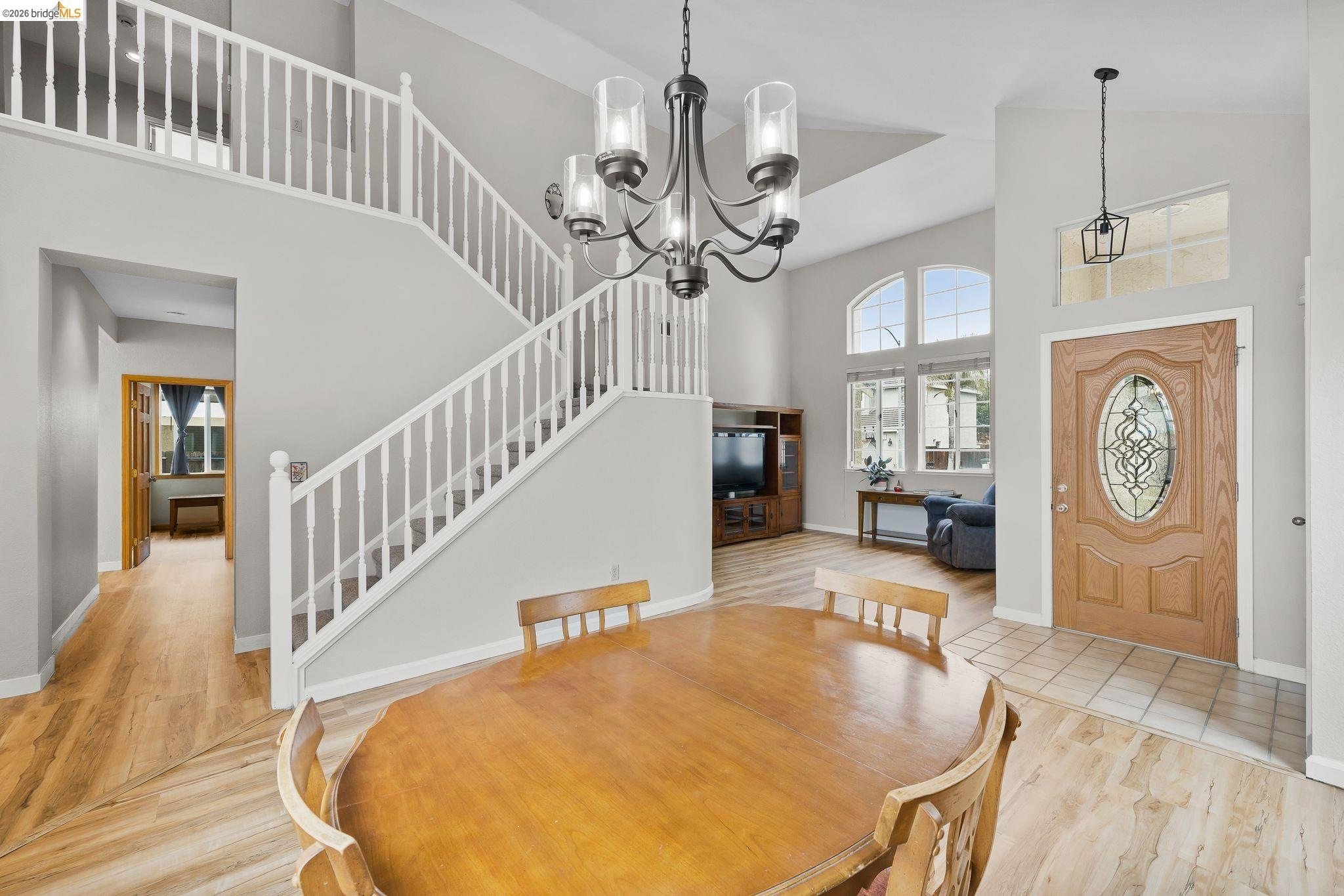 Living room with high ceiling, large windows, and a wooden front door with decorative glass. Provides view of a dining area with a round wooden table and chairs, chandelier, and a staircase leading to the second floor. Features a wood TV stand, couch