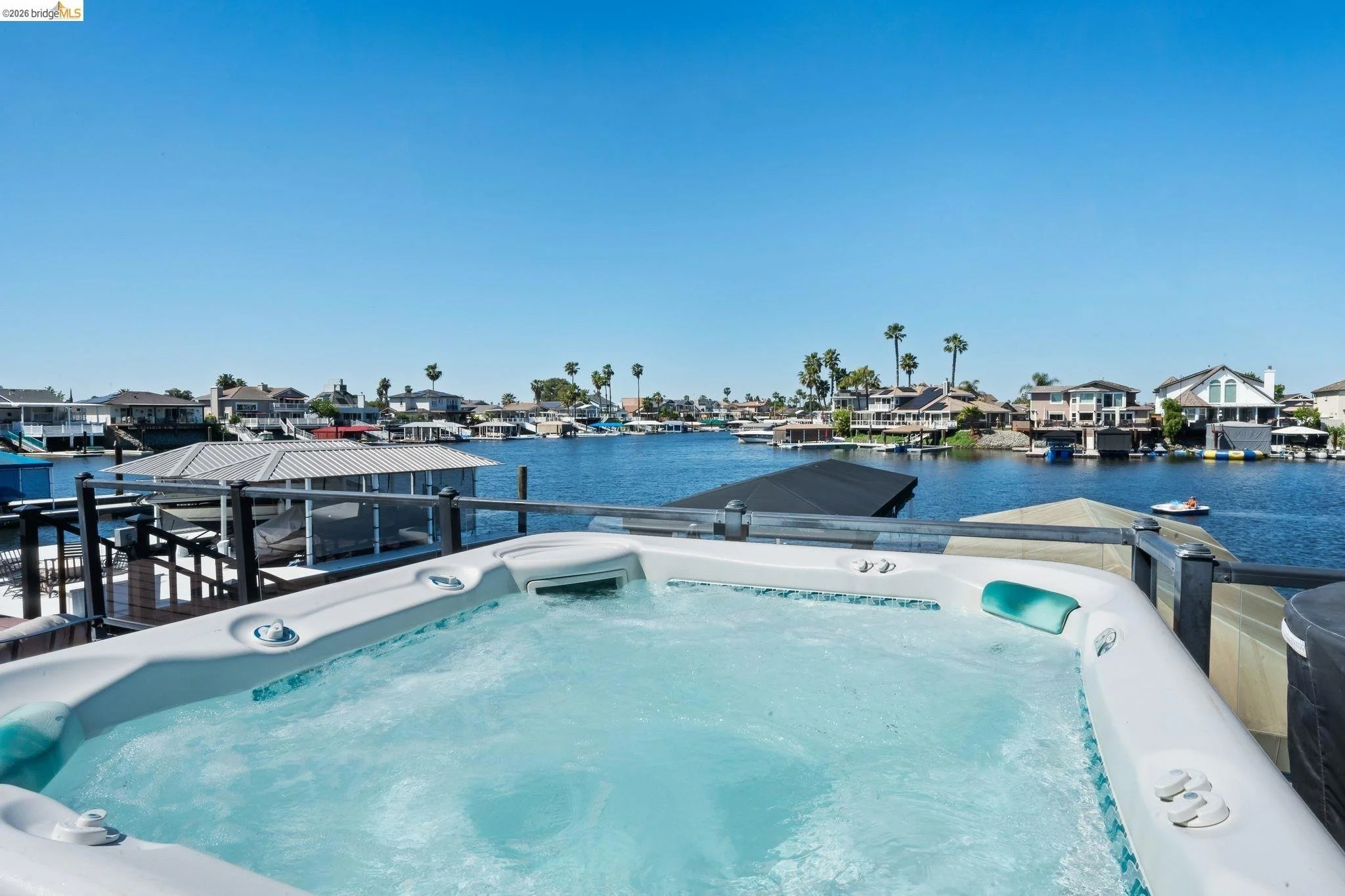 A hot tub on a balcony overlooking a marina with houses, boats, and palm trees on a clear, sunny day.