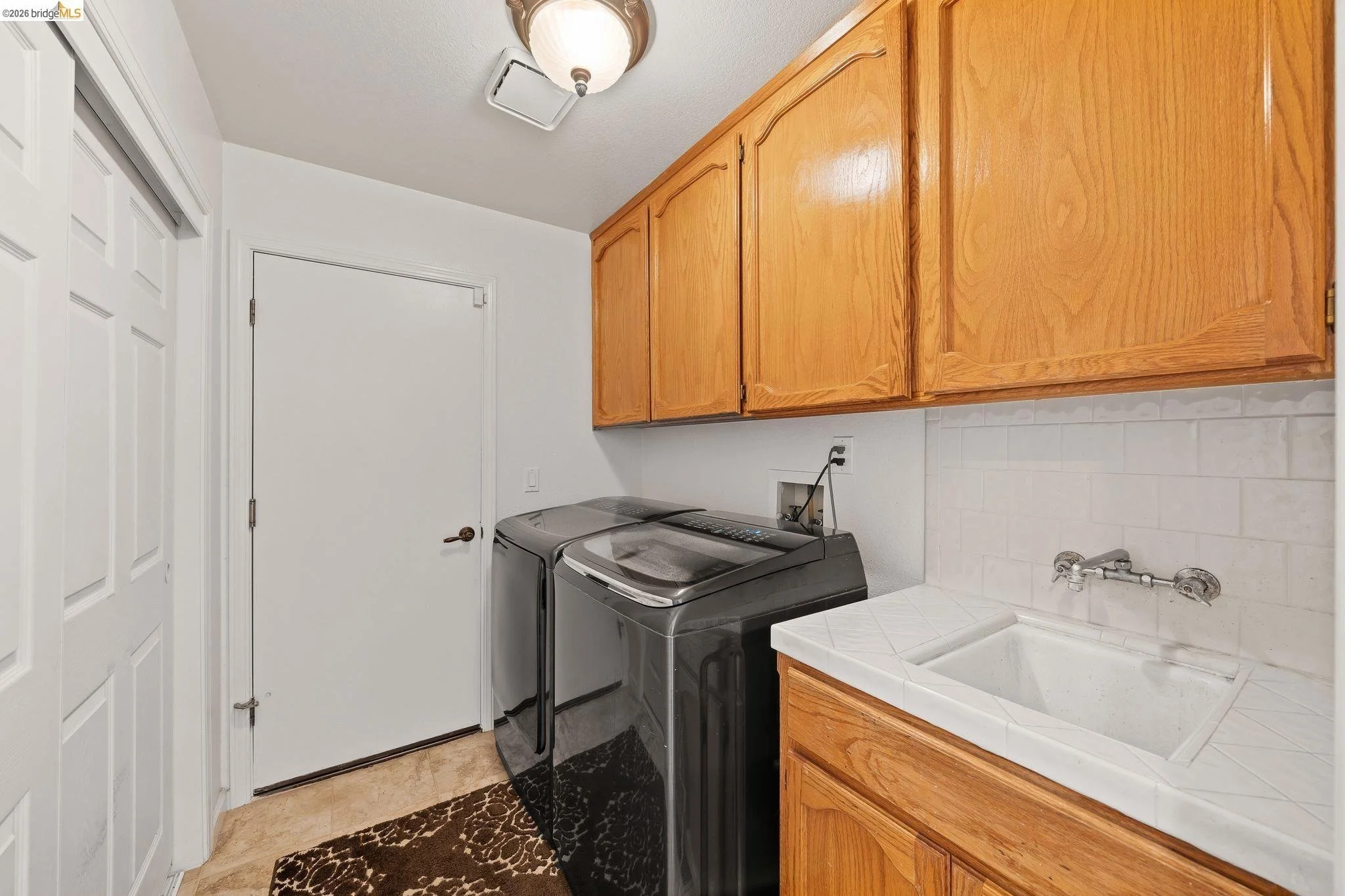 A laundry room with wooden cabinets, a top-loading washing machine and dryer, a utility sink, and a white door. The room has beige tile flooring, a brown rug, and a ceiling light fixture.