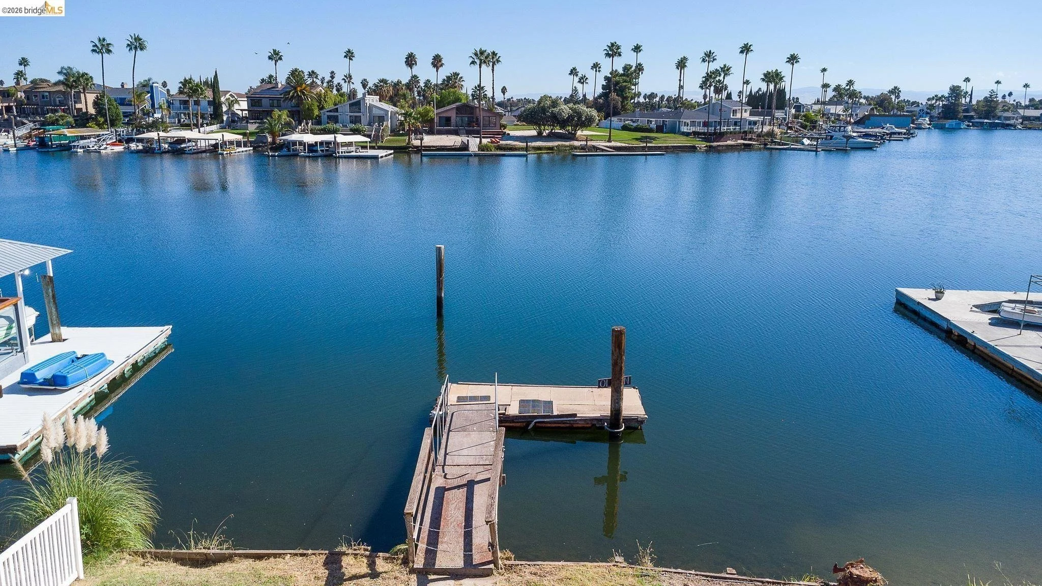 Calm water in a marina with boat docks, houses, and palm trees along the shoreline under a clear blue sky.