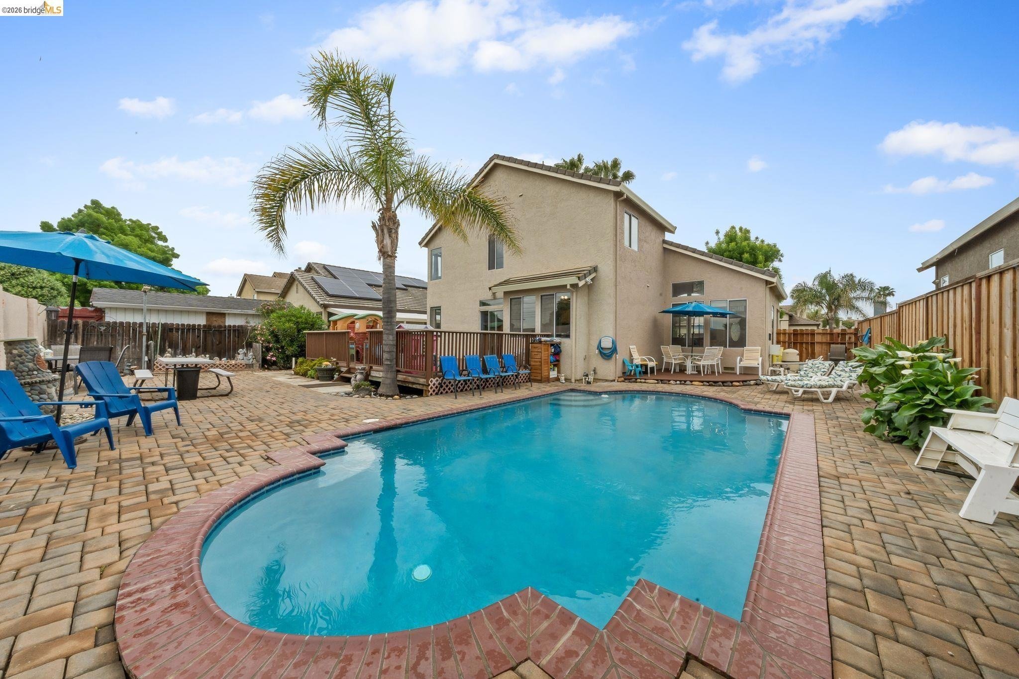 Backyard with a swimming pool, brick patio, lounge chairs, umbrellas, and a two-story house with solar panels and large windows.