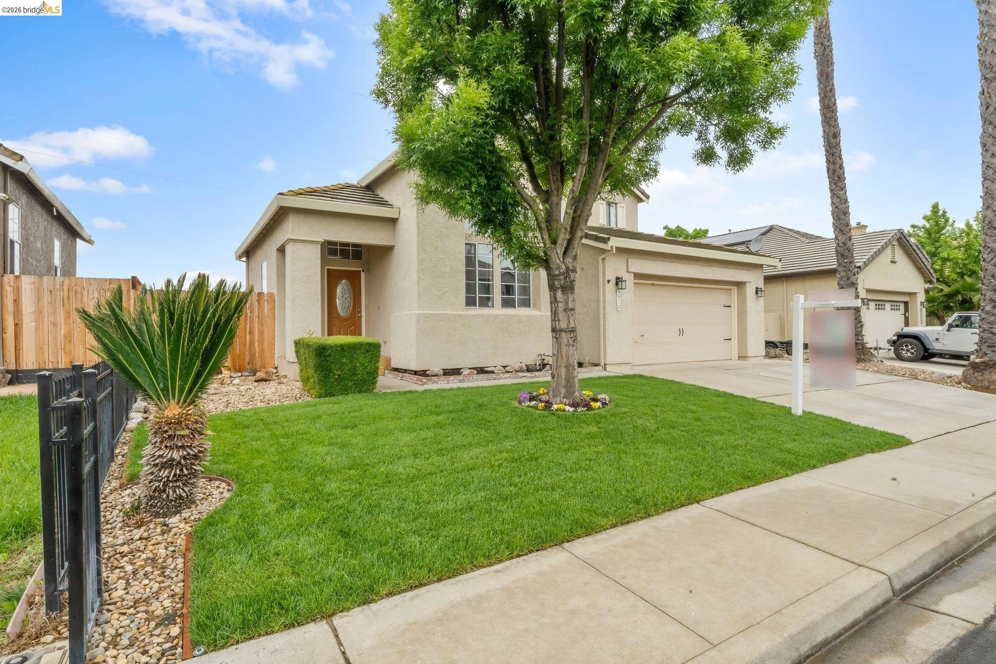 Front view of a single-family house with a well-maintained lawn, a large tree, a small palm, and a flower bed, with neighboring houses and vehicles in the background.