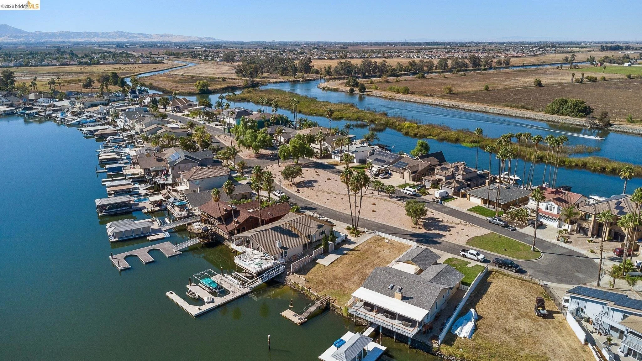 Aerial view of a residential neighborhood with houses along a waterfront canal, boats docked, palm trees, and open land with water in the background.