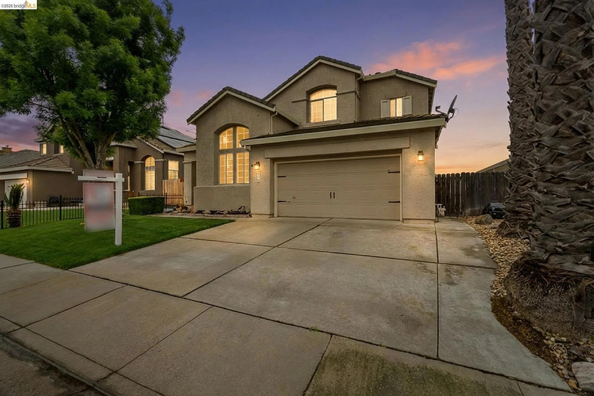 A modern two-story house at sunset with illuminated windows, a two-car garage, a manicured lawn, a large tree, and a palm tree in the front yard.