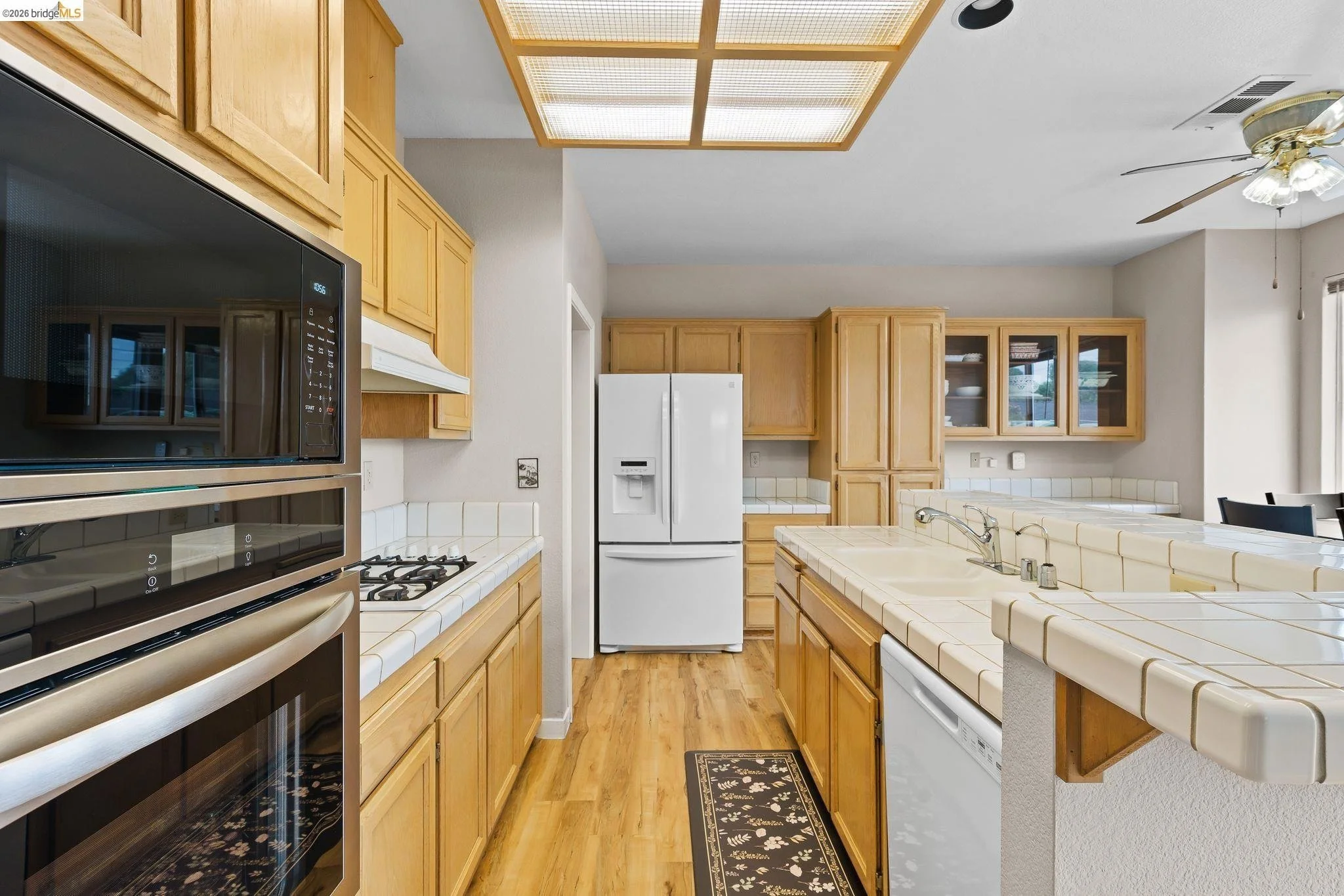 Kitchen with wooden cabinets, white appliances including a refrigerator, oven, microwave, and dishwasher. There is a tiled countertop with a sink, a ceiling fan, and a wooden light fixture on the ceiling. Hardwood flooring and a small decorative rug 