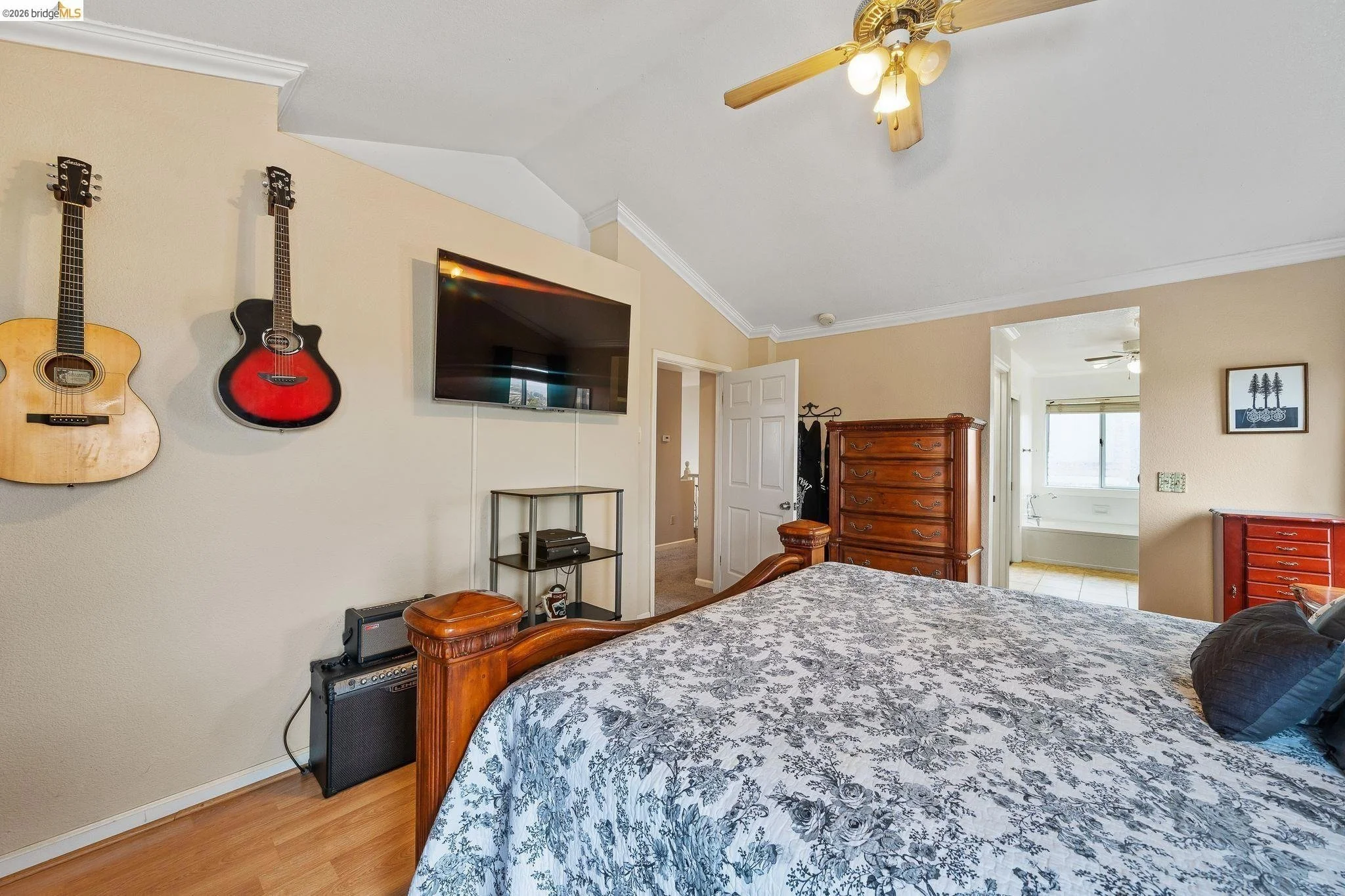 Bedroom with wooden bed frame, floral bedspread, guitars on the wall, flat-screen TV, chest of drawers, and a view of a bathroom with a bathtub through an open door.