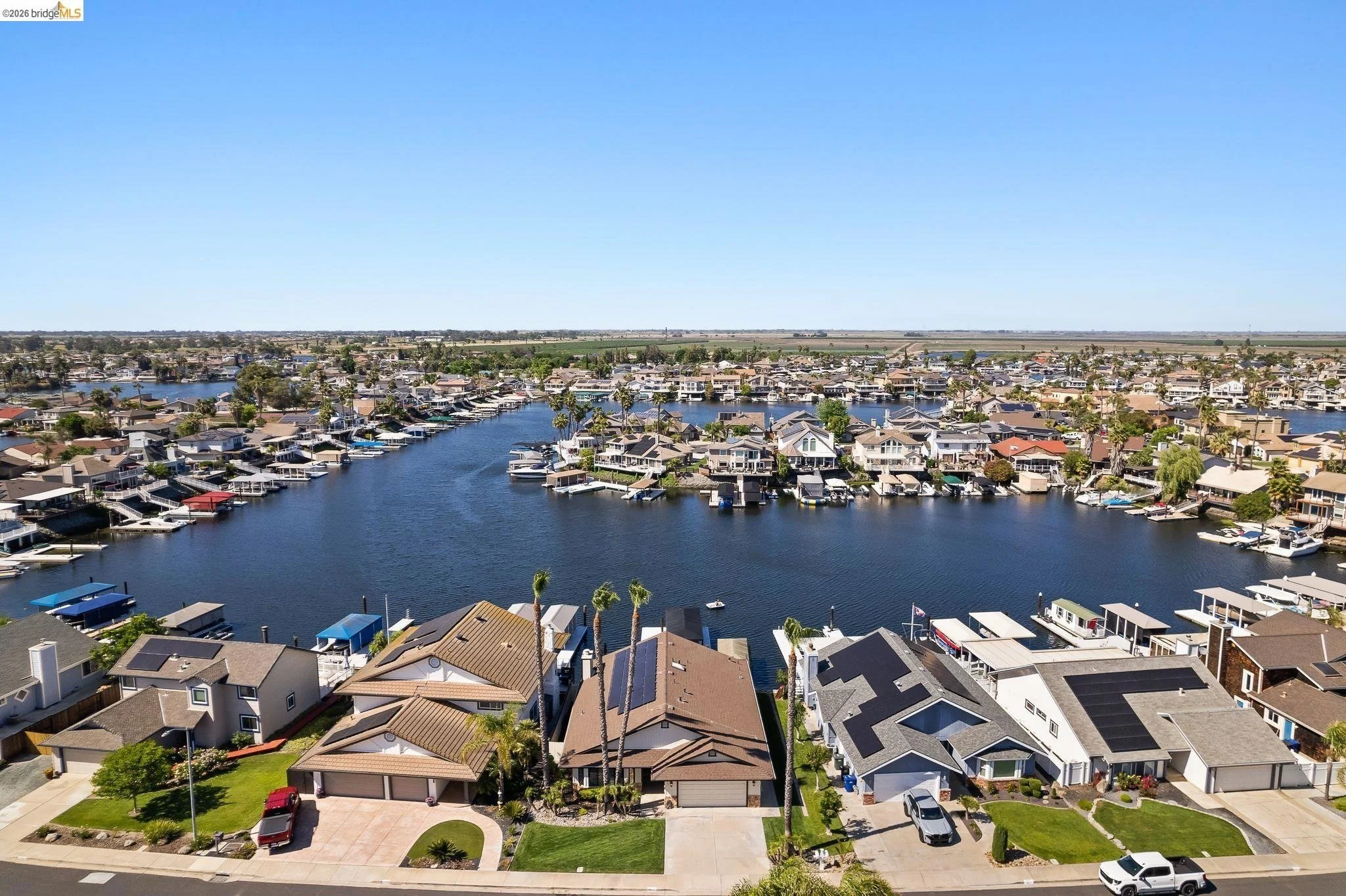 Aerial view of a residential neighborhood with houses along a waterway, boats docked at piers, and clear blue sky.