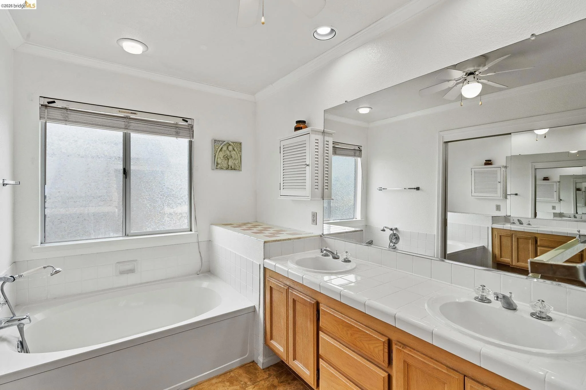 A clean bathroom with a large mirror, two sinks with wooden cabinets, a bathtub under two windows, white tile surfaces, a ceiling fan, and frosted windows for privacy.