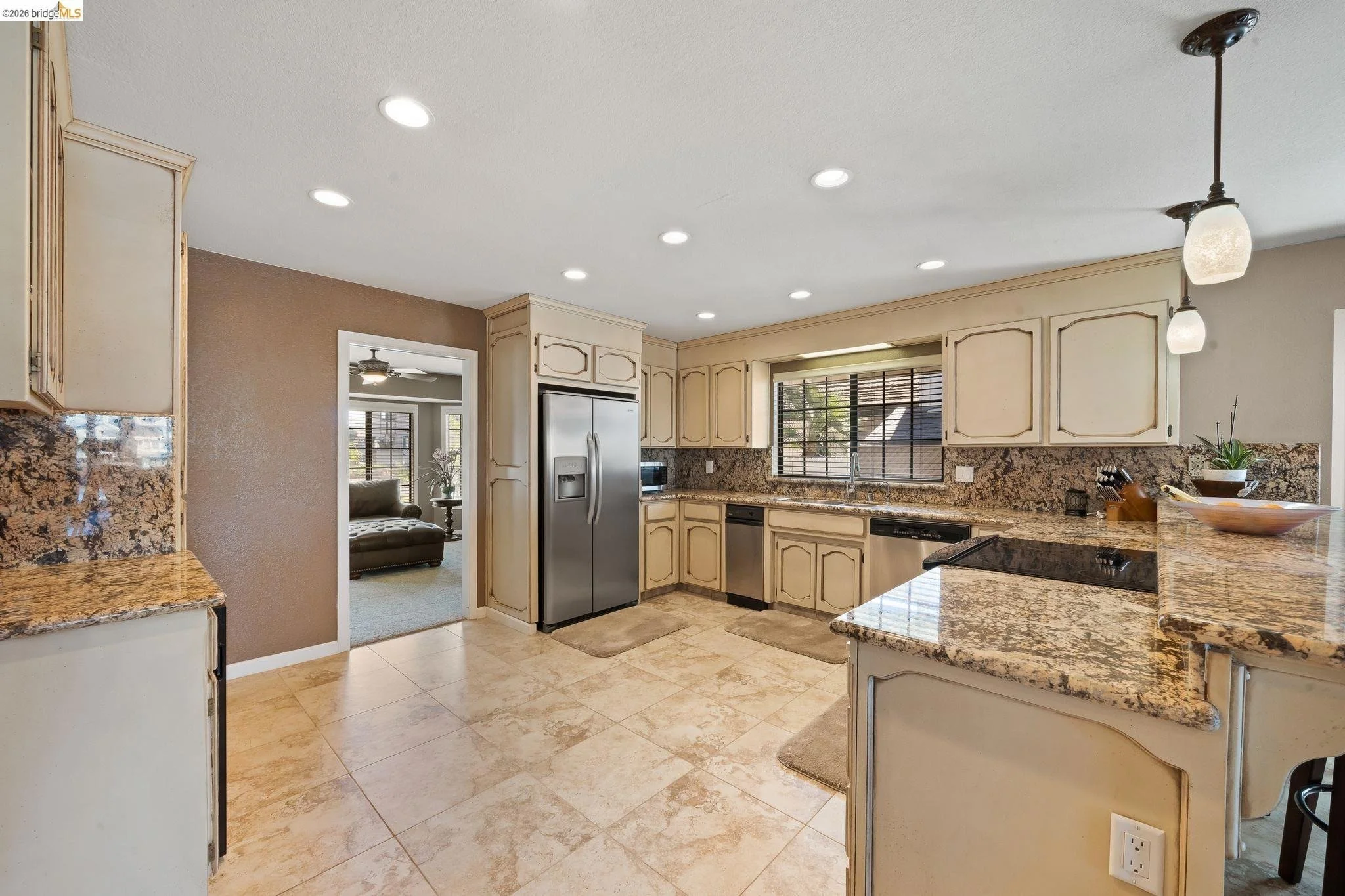 Kitchen with beige cabinets, granite countertops, stainless steel refrigerator, dishwasher, and stove, beige tiled floor, window with blinds, and pendant lights.
