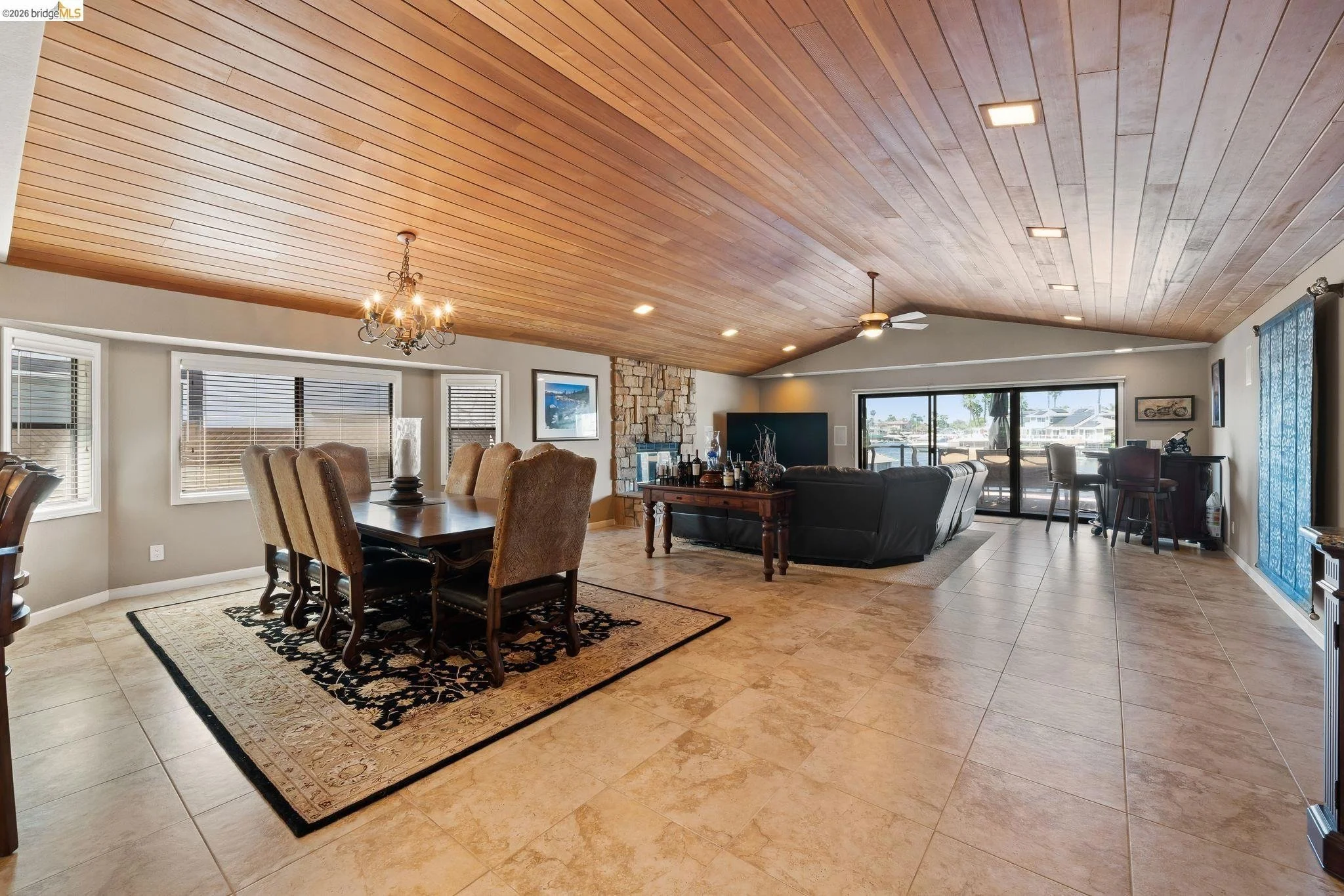 Open living and dining room with a wooden ceiling, chandelier, and ceiling fan. The dining area has a large wooden table with eight upholstered chairs on a patterned rug. The living area features a black leather sofa, TV, stone fireplace, and sliding