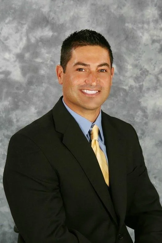 A man in a black suit, light blue shirt, and yellow tie, smiling in front of a gray mottled background.