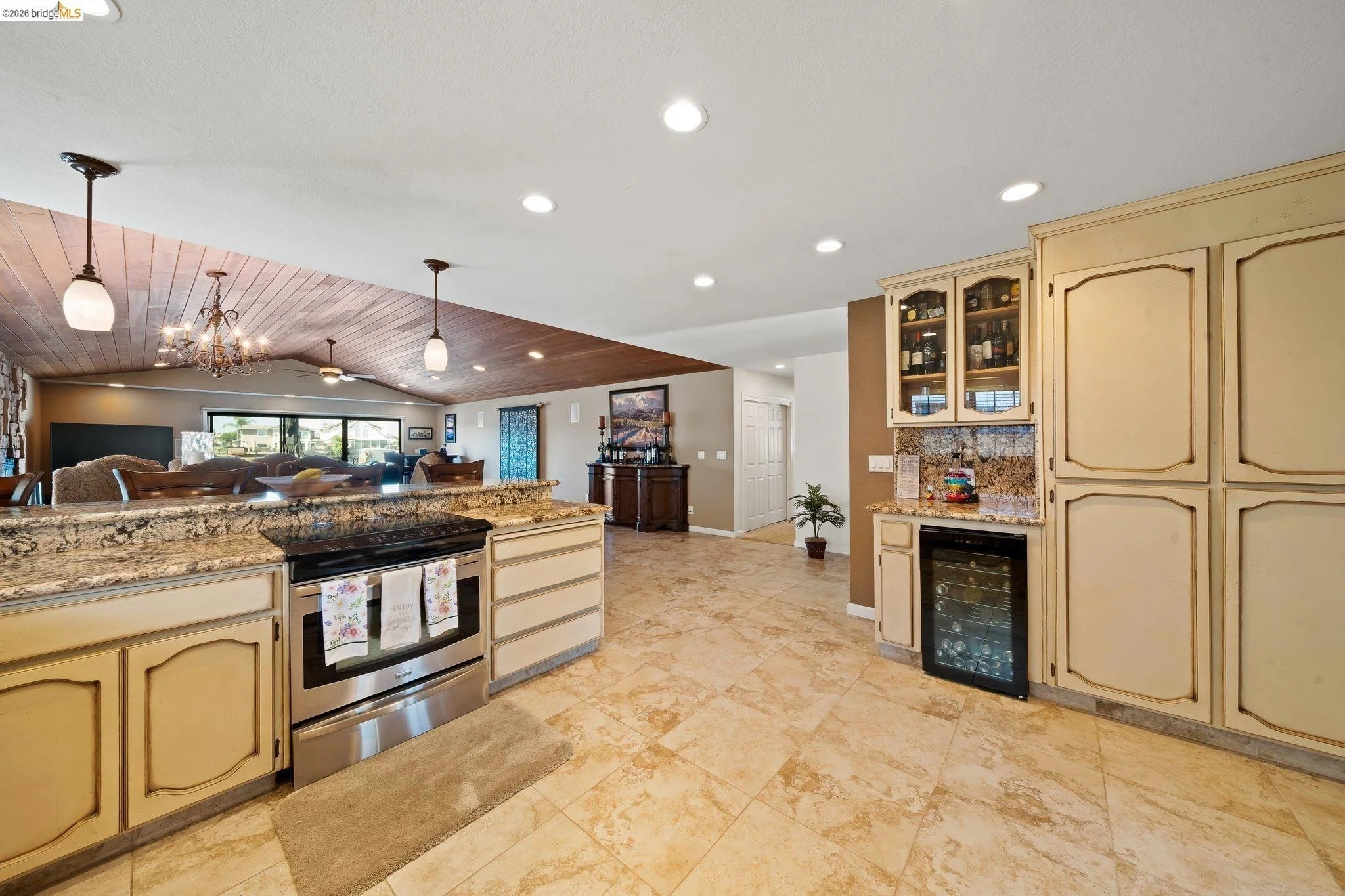 Open kitchen with beige cabinets, granite countertops, stainless steel oven, and a wine cooler. The floor is tile, and the ceiling has recessed lighting. The kitchen opens to a living area with a wood-paneled vaulted ceiling, a chandelier, and large 