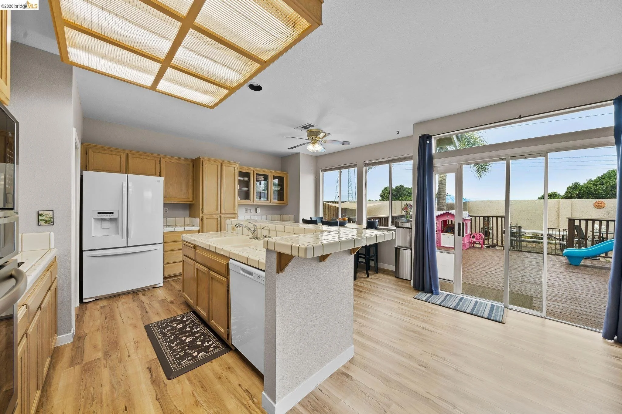 Kitchen with wooden cabinets, white appliances, tile countertops, and a sliding glass door leading to a deck with outdoor play equipment.