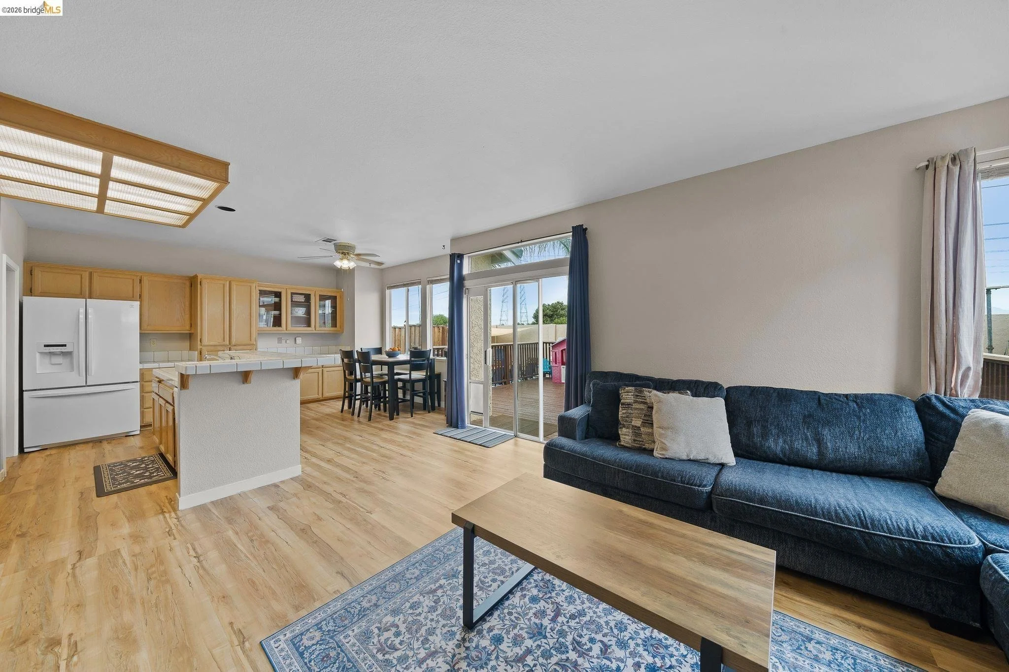 Open-concept living room and kitchen in a modern home with wood floors, a dark blue sofa, a wooden coffee table, and large windows with curtains.